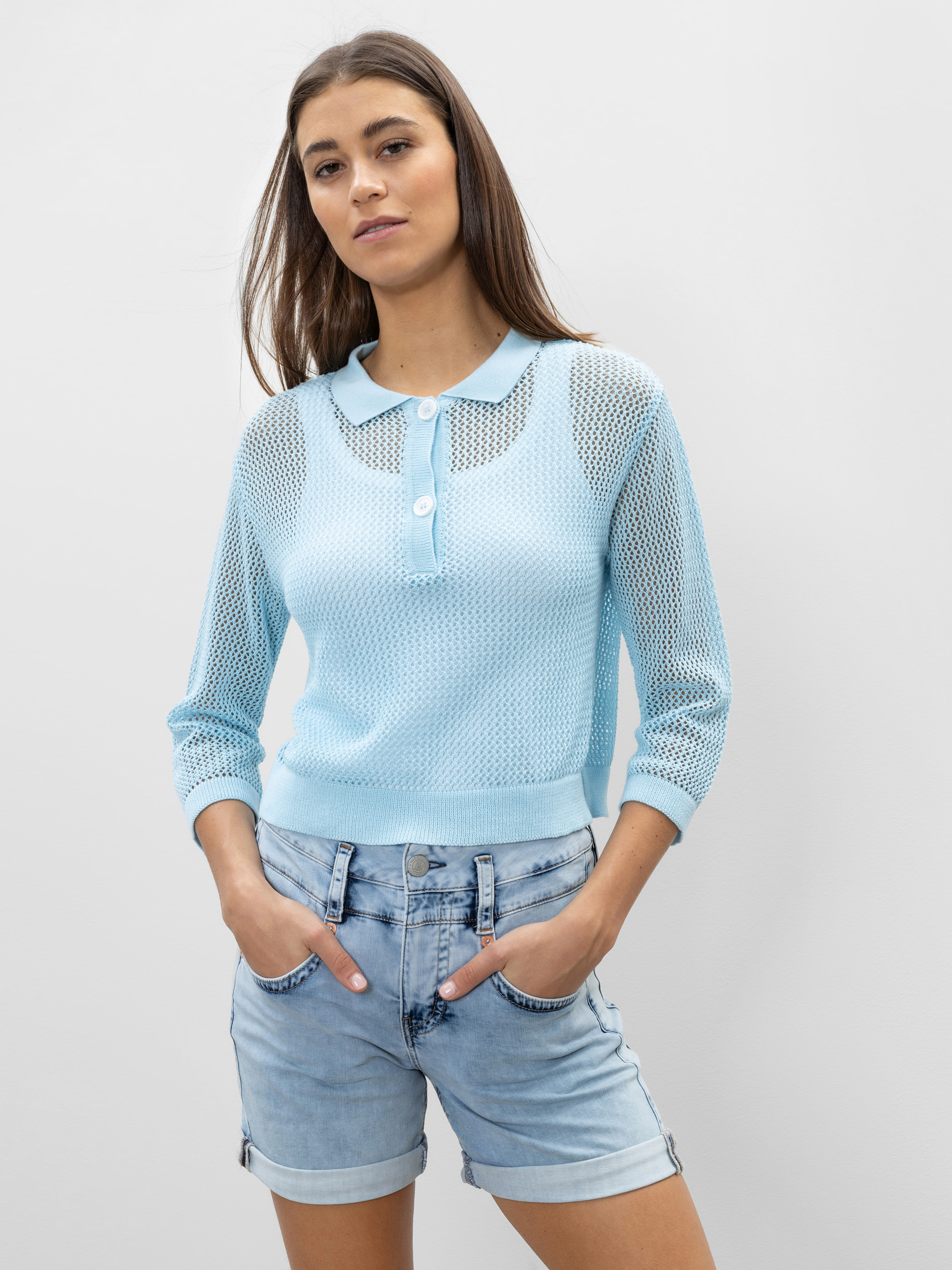 Eine Frau trägt einen hellblauen, langärmeligen Netzstrickpullover mit Kragen und Knöpfen, kombiniert mit hellen Jeansshorts. Sie steht mit den Händen in den Taschen vor einem schlichten hellen Hintergrund.