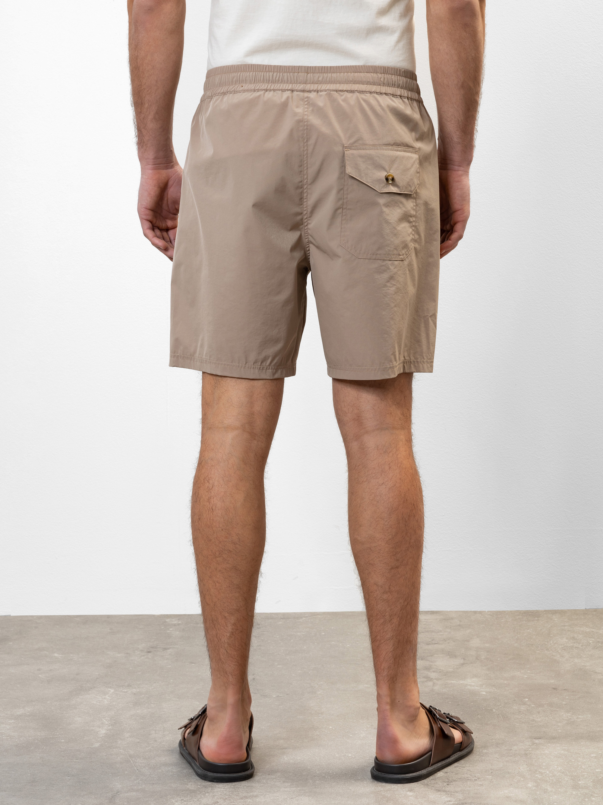 Ein Mann in hellbraunen Shorts mit Gesäßtasche und braunen Sandalen steht auf einem grauen Boden und schaut von der Kamera weg. Der Hintergrund ist einfarbig weiß.