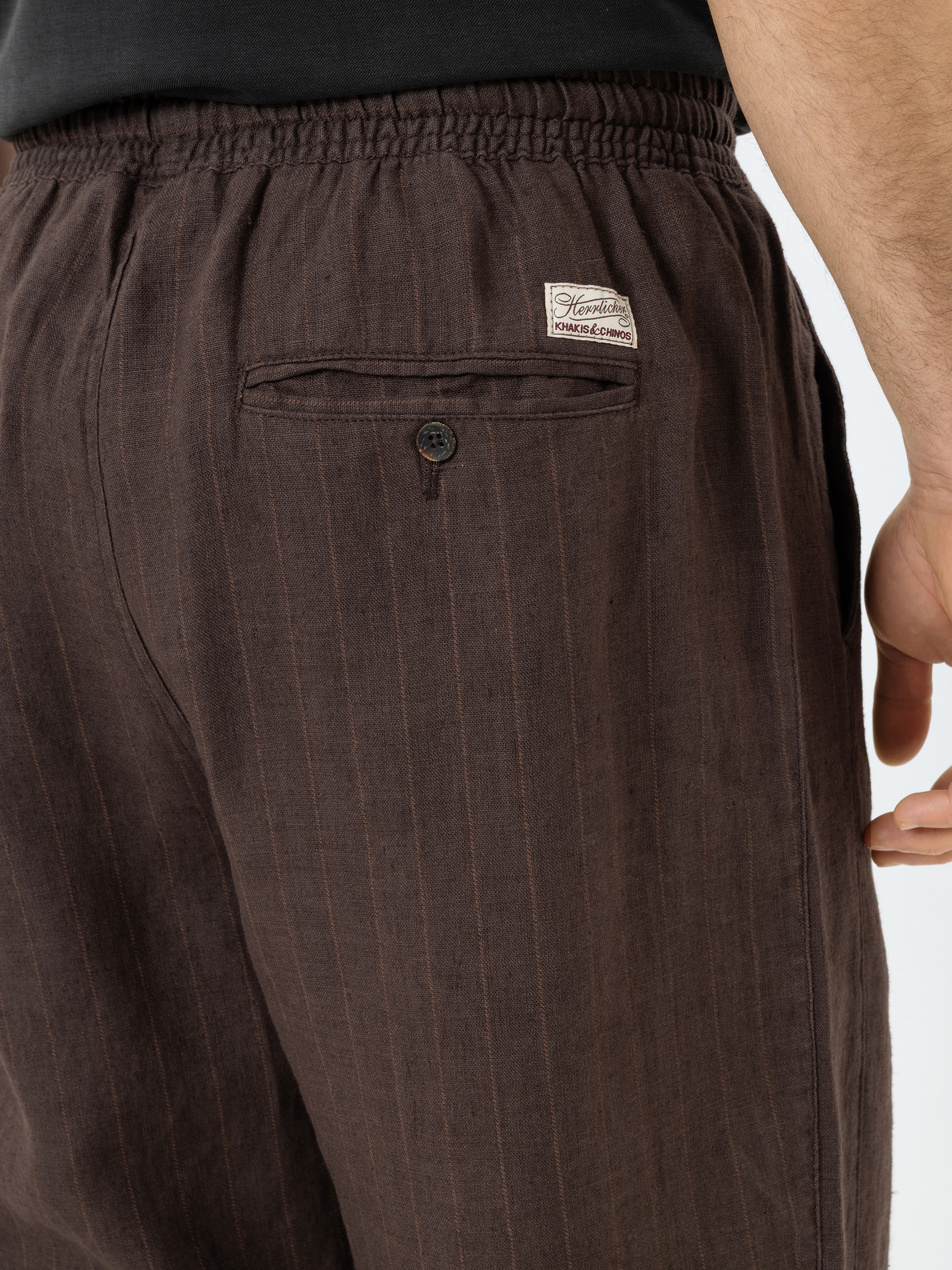 Nahaufnahme einer Person, die eine braune, gestreifte Hose mit einem elastischen Bund, einer Gesäßtasche mit Knopf und einem kleinen Etikett über der Tasche trägt. Die rechte Hand der Person ist an ihrer Seite zu sehen.