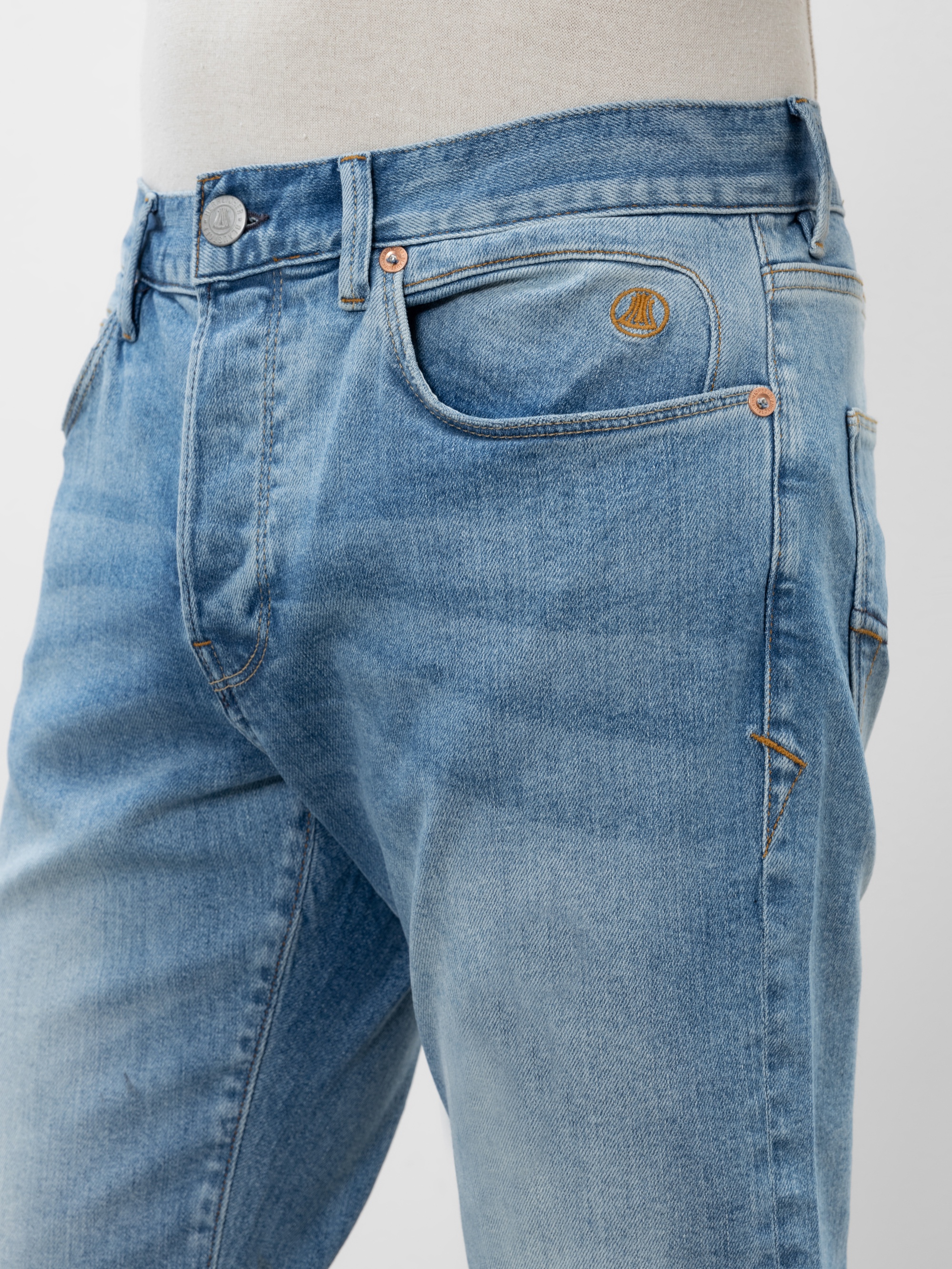 Nahaufnahme einer Person, die eine hellblaue Jeans mit orangefarbenen Nähten, einem Metallknopf und einem kleinen gestickten Logo über der Vordertasche trägt. Der Hintergrund ist schlicht und neutral.