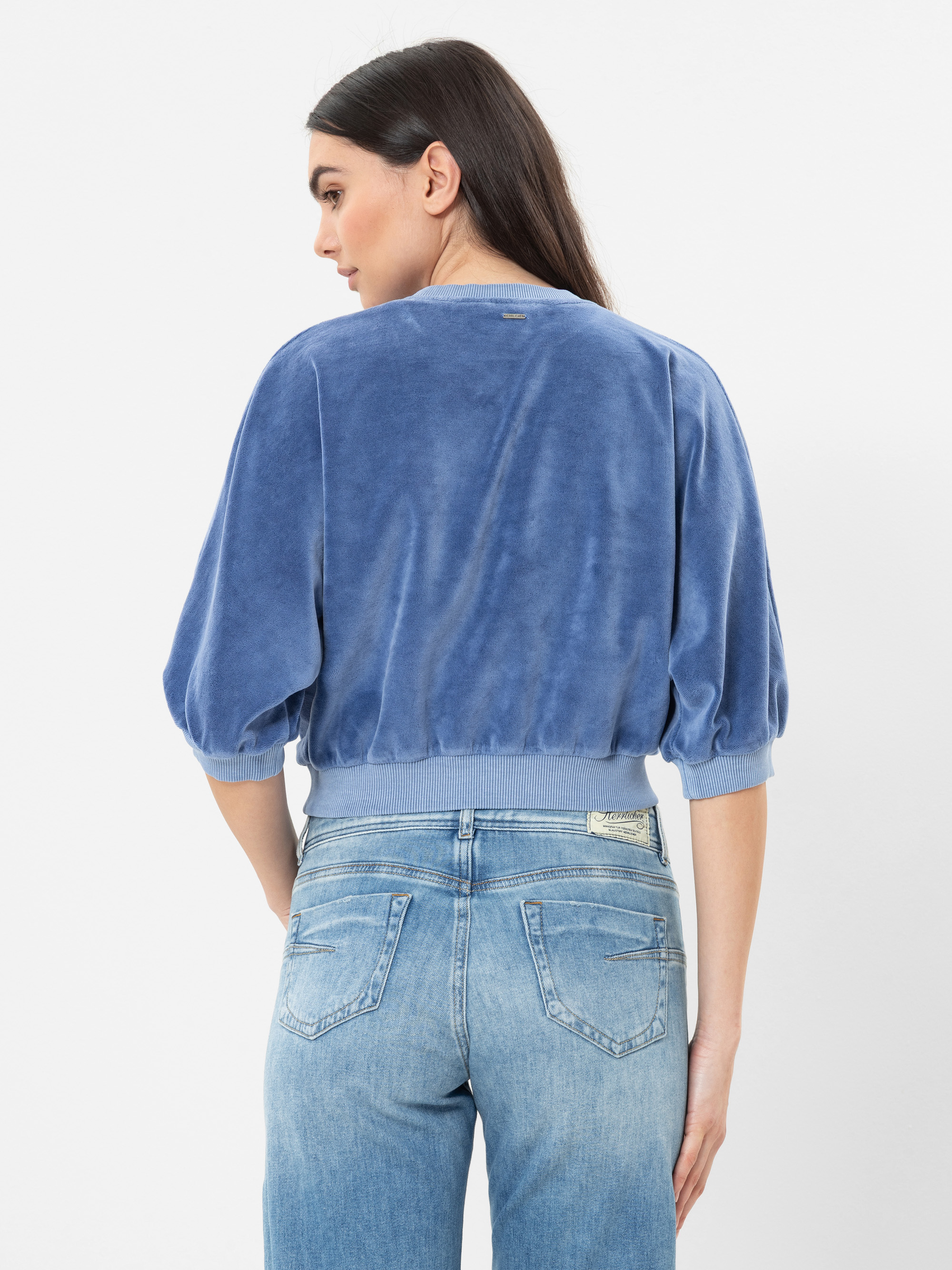 Eine Frau mit langen dunklen Haaren trägt ein blaues, locker sitzendes Veloursoberteil mit Dreiviertelärmeln und hellblaue Jeans mit hoher Taille. Sie blickt von sich weg über ihre Schulter auf einen schlichten weißen Hintergrund.