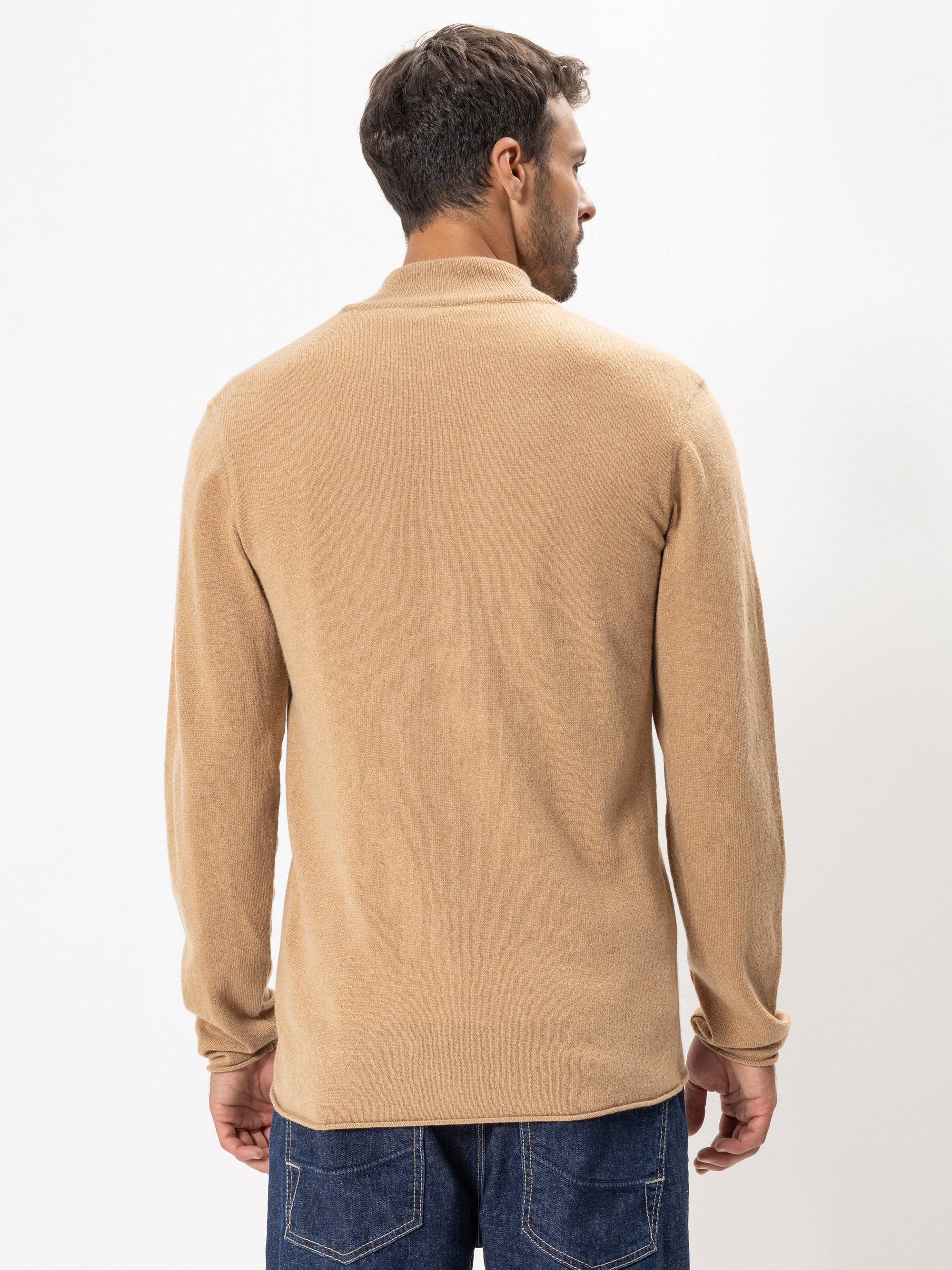 Ein Mann steht mit dem Gesicht zur Kamera, trägt einen hellbraunen langärmeligen Pullover und dunkelblaue Jeans vor einem schlichten weißen Hintergrund.