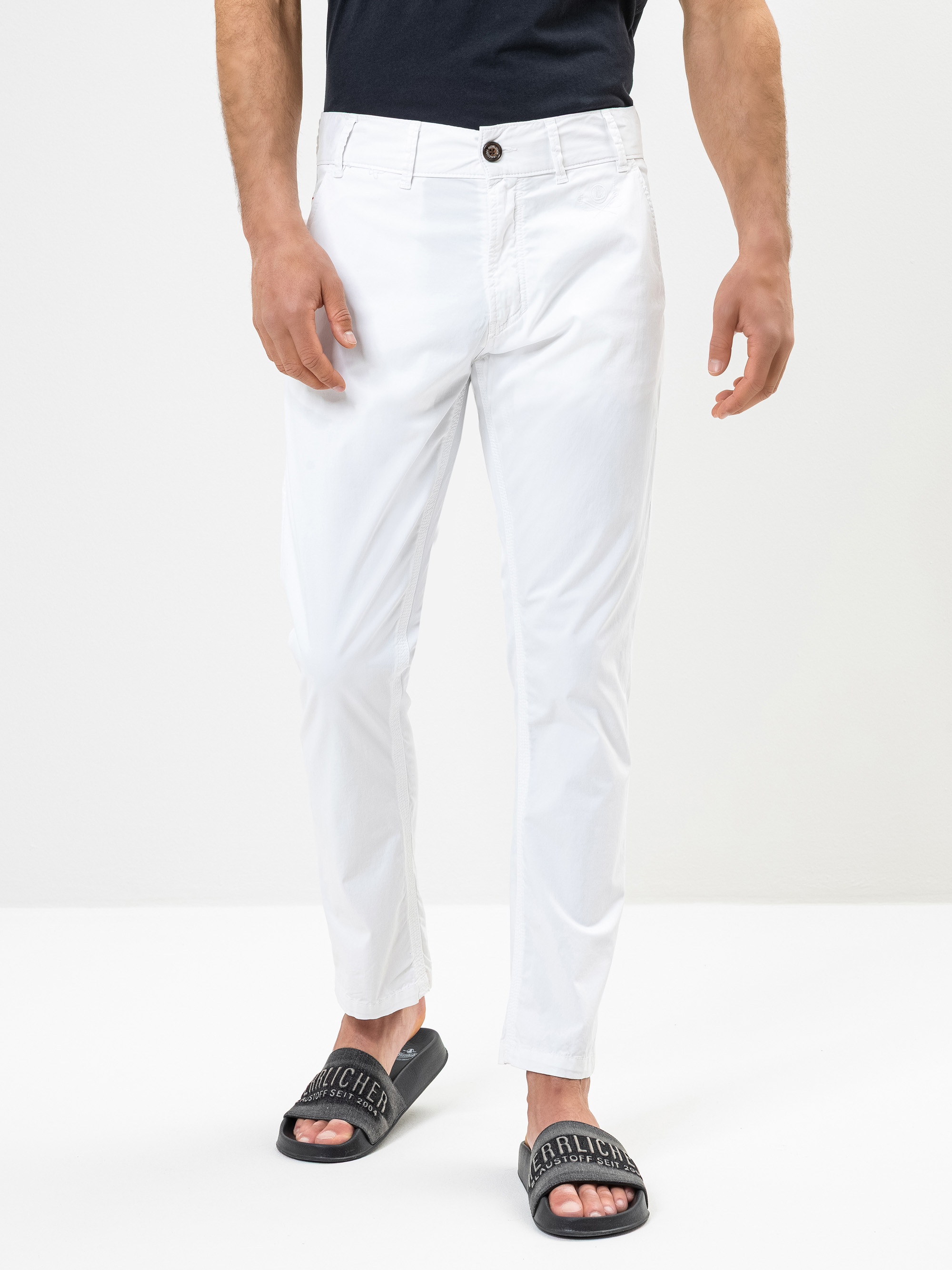 Ein Mann, der ein schwarzes T-Shirt, eine weiße Hose und schwarze Slipper-Sandalen trägt, steht vor einem schlichten weißen Hintergrund. Das Bild ist an den Schultern abgeschnitten und konzentriert sich auf die Hose und die Sandalen.