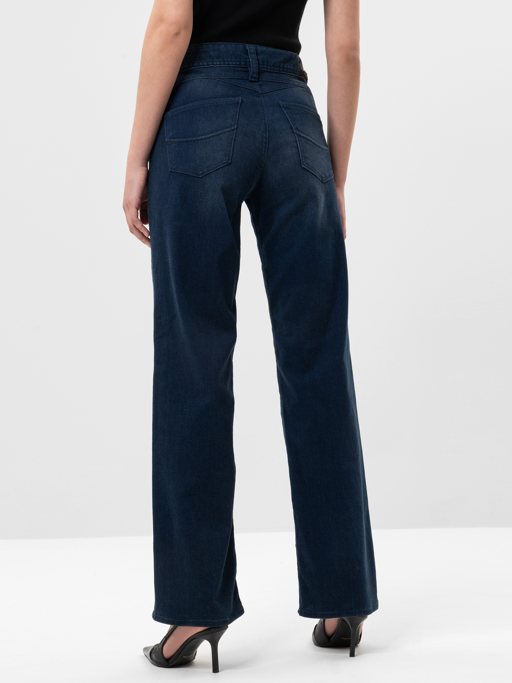 Eine Person, die dunkelblaue Jeans mit weitem Bein und schwarze Schuhe mit Absätzen trägt, steht mit dem Gesicht zur Kamera vor einem schlichten hellen Hintergrund.