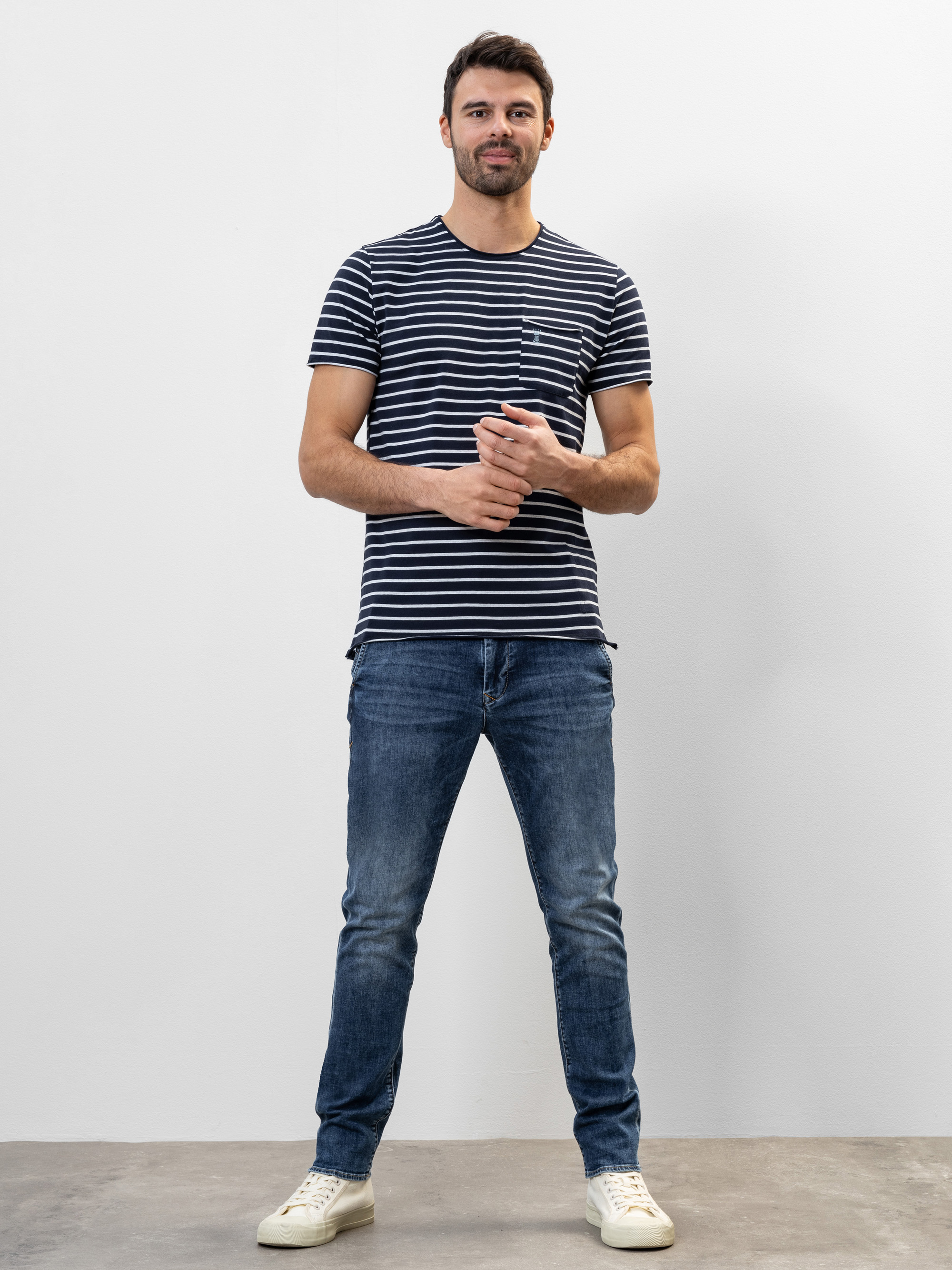 Ein Mann steht vor einer schlichten weißen Wand und trägt ein marineblau-weiß gestreiftes T-Shirt, blaue Jeans und weiße Turnschuhe. Er hat kurzes dunkles Haar, einen Bart und lächelt sanft, während er die Hände locker verschränkt.