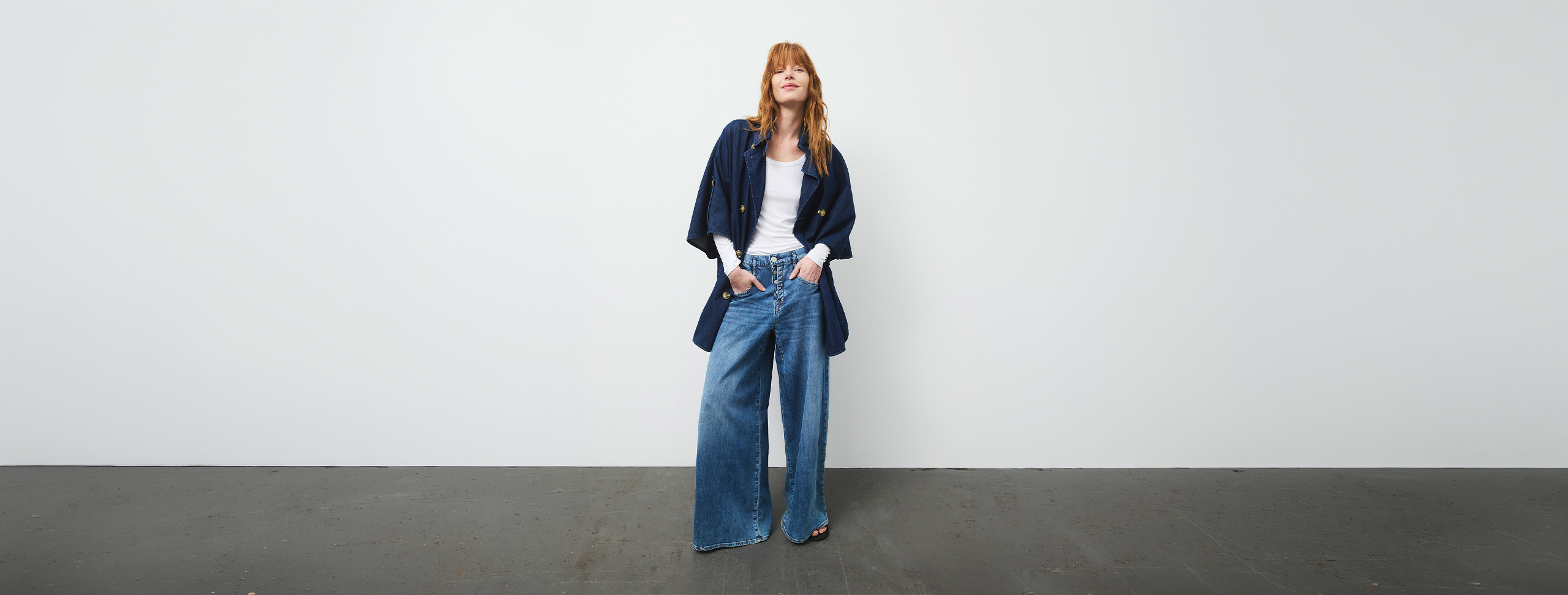 Eine Person steht vor einer schlichten weißen Wand, trägt eine marineblaue Jacke, ein weißes Oberteil und eine weitbeinige Bluejeans, die Hände in den Taschen und einen entspannten Gesichtsausdruck.