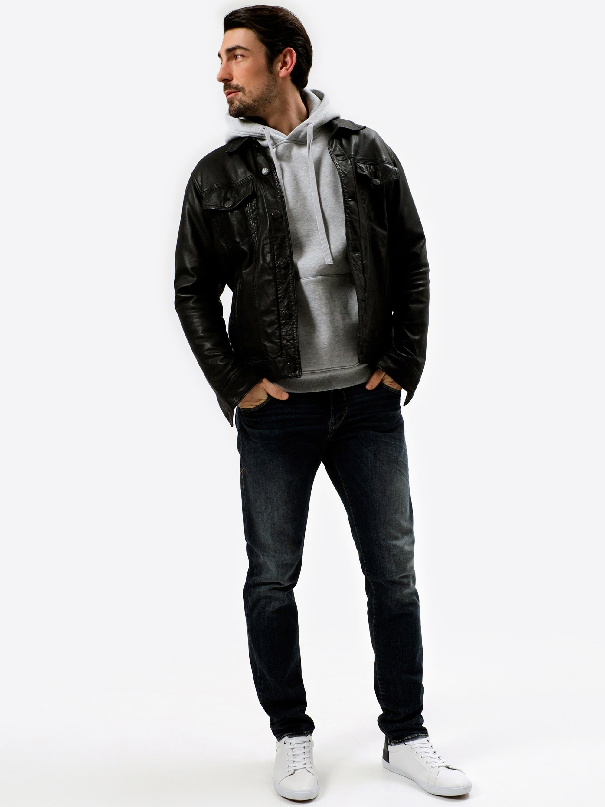 Ein bärtiger Mann steht mit den Händen in den Jackentaschen, trägt eine schwarze Lederjacke über einem grauen Kapuzenpulli, dunkle Jeans und weiße Turnschuhe und blickt zur Seite vor einem schlichten weißen Hintergrund.
