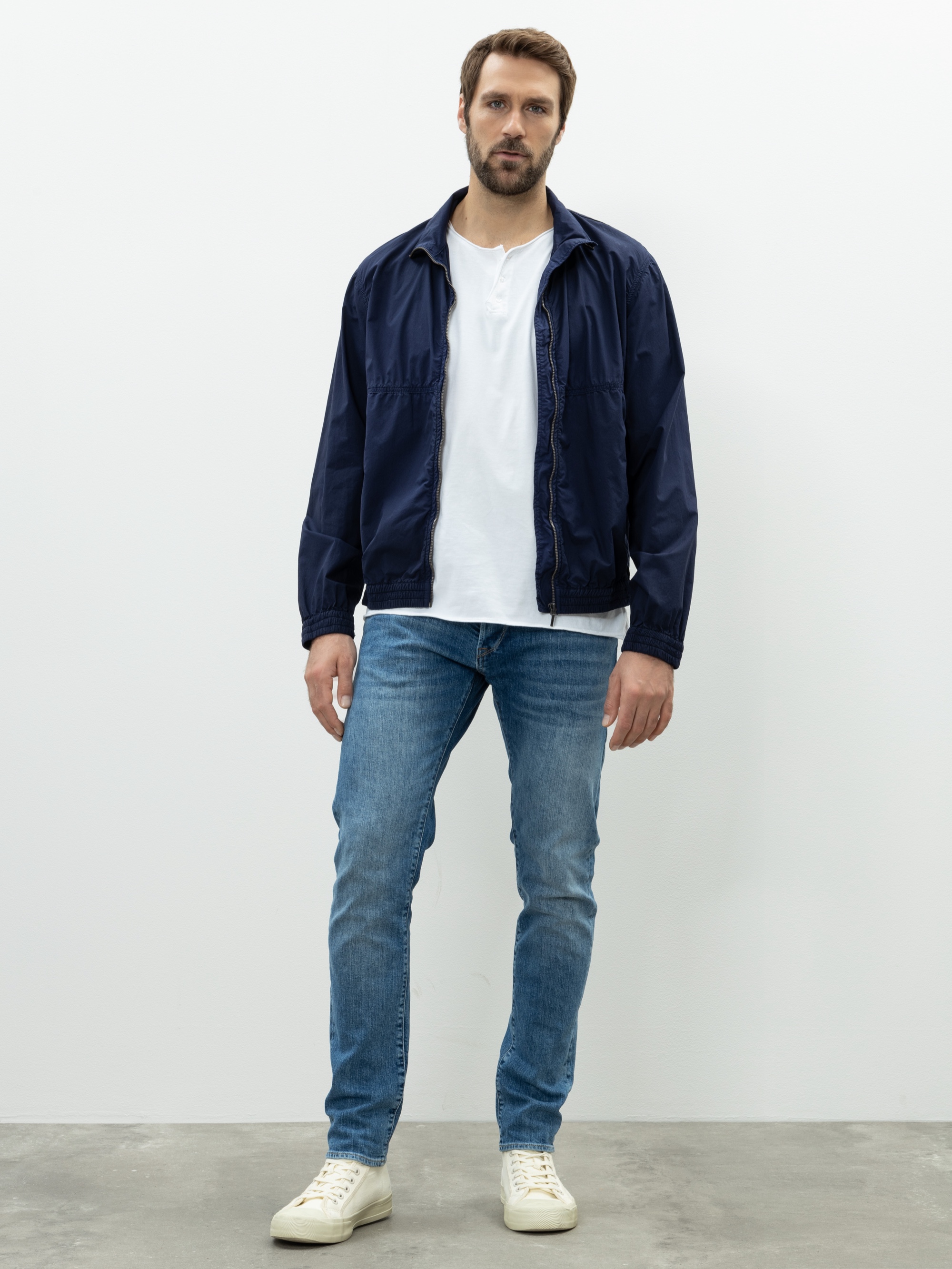 Ein Mann mit kurzen braunen Haaren und einem Bart trägt eine marineblaue Jacke über einem weißen Hemd, blaue Jeans und weiße Turnschuhe und steht vor einem schlichten weißen Hintergrund.