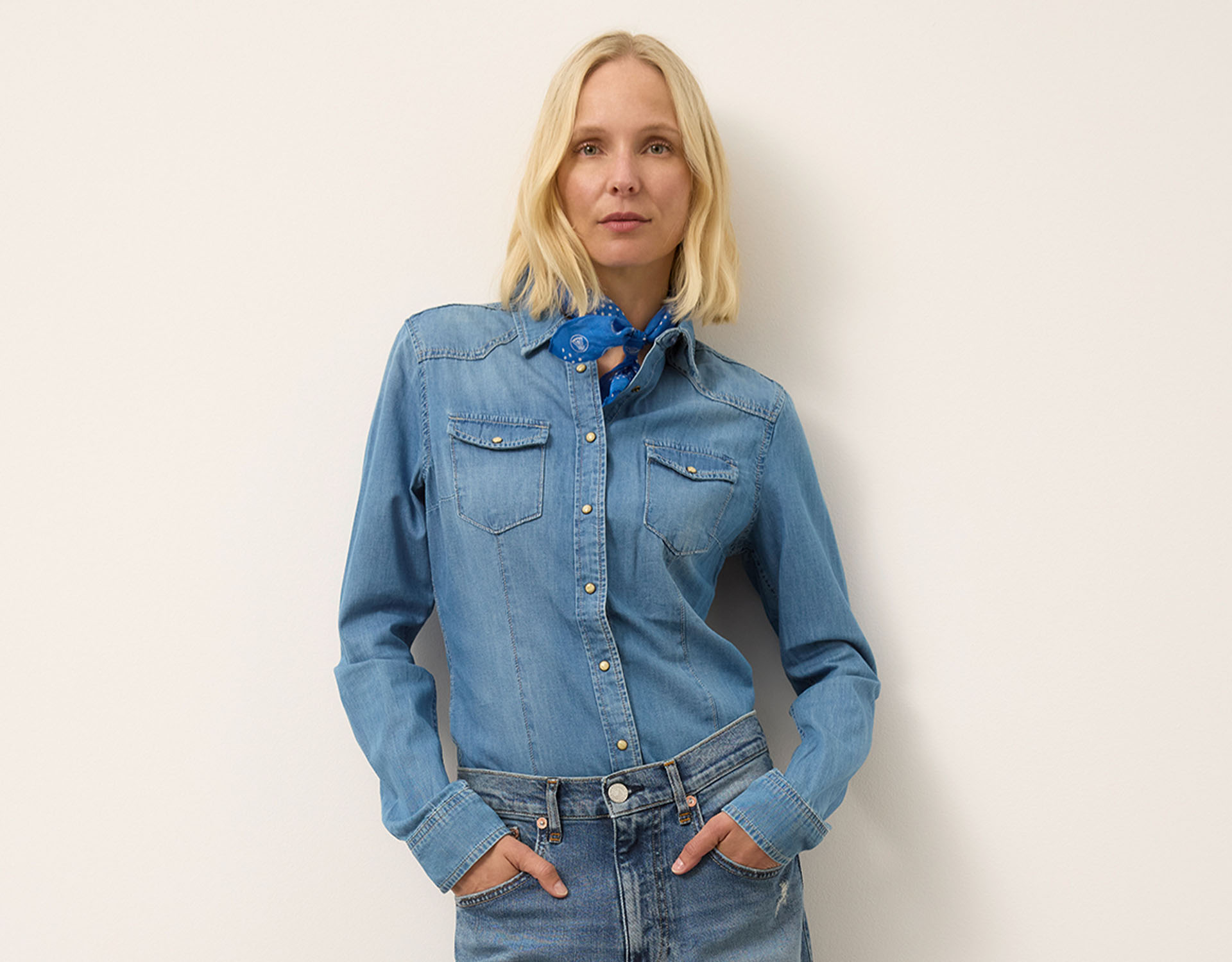 Eine Person mit blonden Haaren, die ein hellblaues Jeanshemd, blaue Jeans und einen blauen Schal trägt, steht mit den Händen in den Taschen vor einer schlichten hellen Wand.