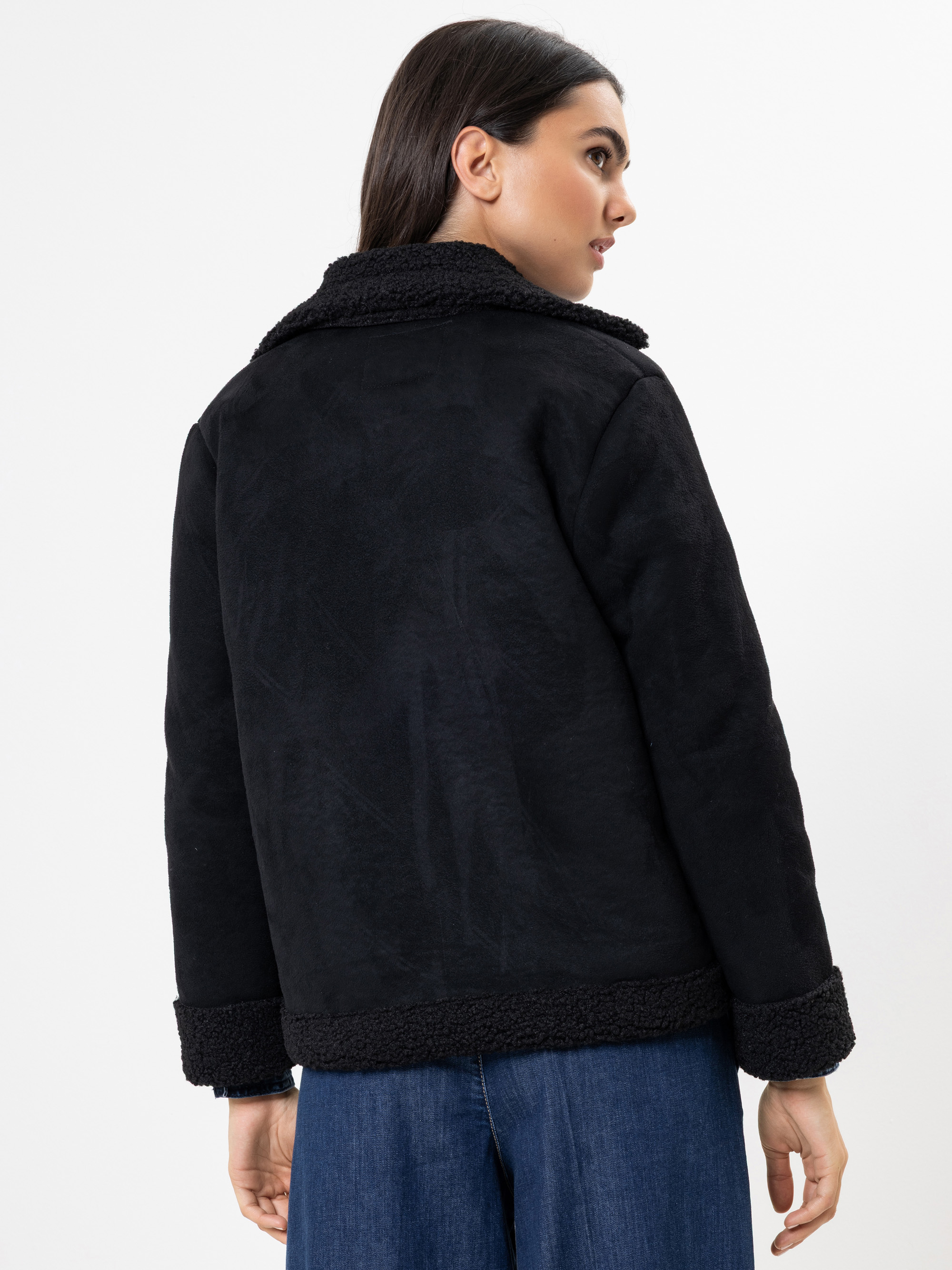 Eine Frau mit langen dunklen Haaren wird von hinten gezeigt. Sie trägt eine schwarze Shearling-Jacke mit breitem Kragen und Manschettenärmeln. Sie trägt außerdem eine blaue Jeans und steht vor einem schlichten weißen Hintergrund.