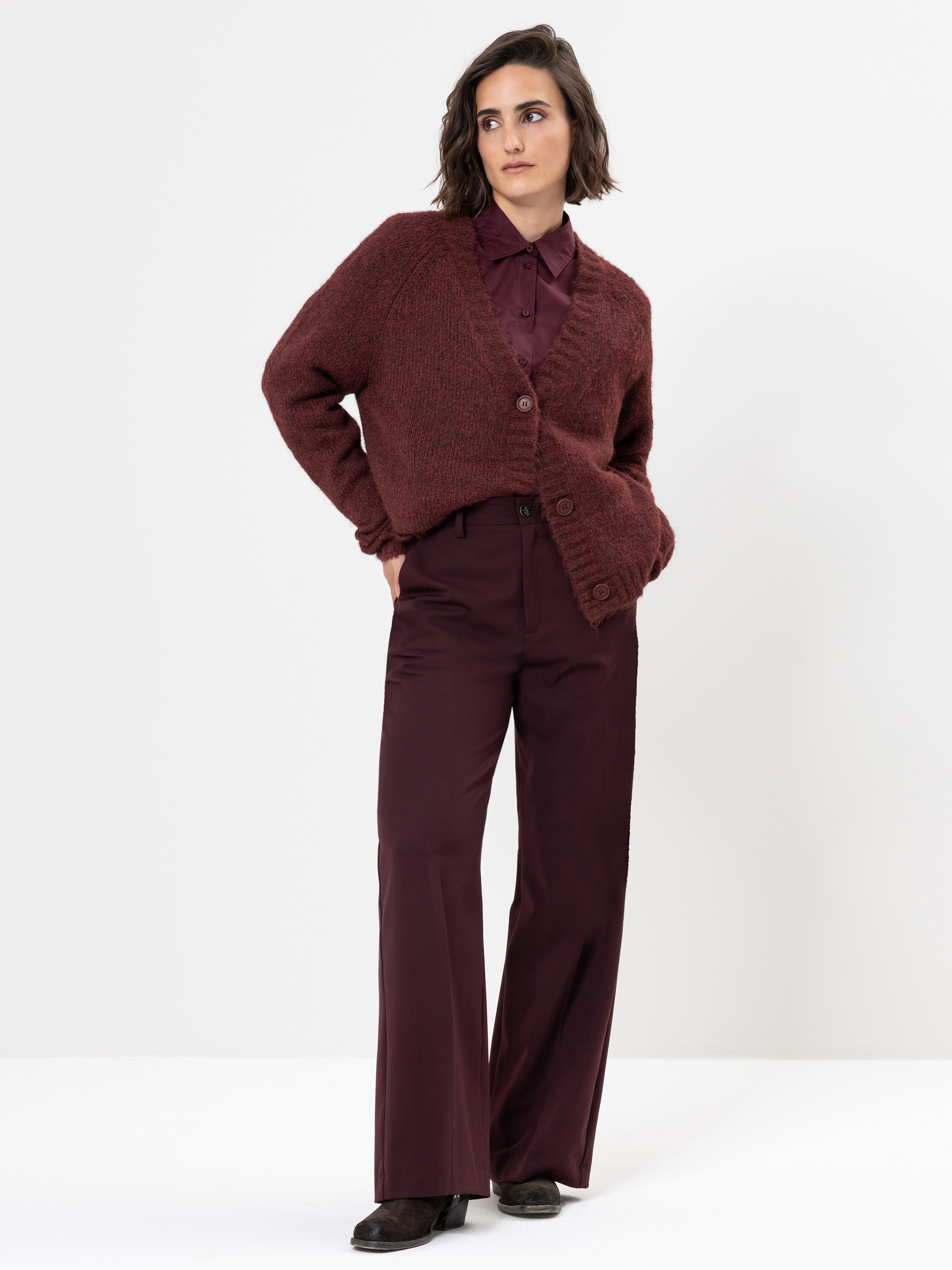 Eine Person steht vor einem weißen Hintergrund und trägt eine kastanienbraune Strickjacke über einem kastanienbraunen Button-up-Hemd, eine weit geschnittene kastanienbraune Hose und dunkle Schuhe. Sie hat kurzes, gewelltes Haar und einen neutralen Gesi