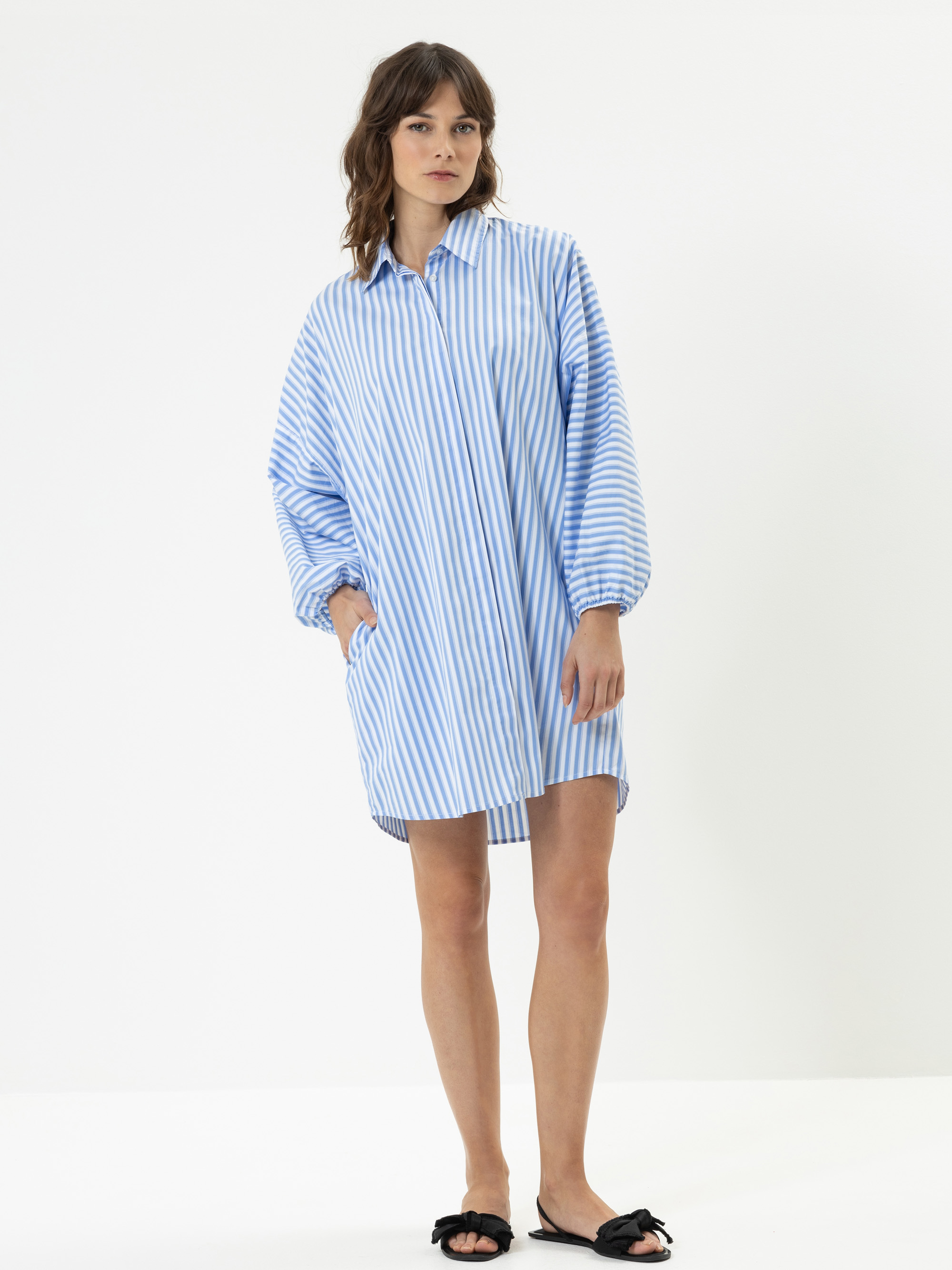 Eine Frau steht vor einem schlichten weißen Hintergrund und trägt ein locker sitzendes, langärmeliges, blau-weißes, vertikal gestreiftes Hemdkleid und schwarze Slide-Sandalen mit Schleife. Sie hat gewelltes Haar und einen neutralen Gesichtsausdruck.