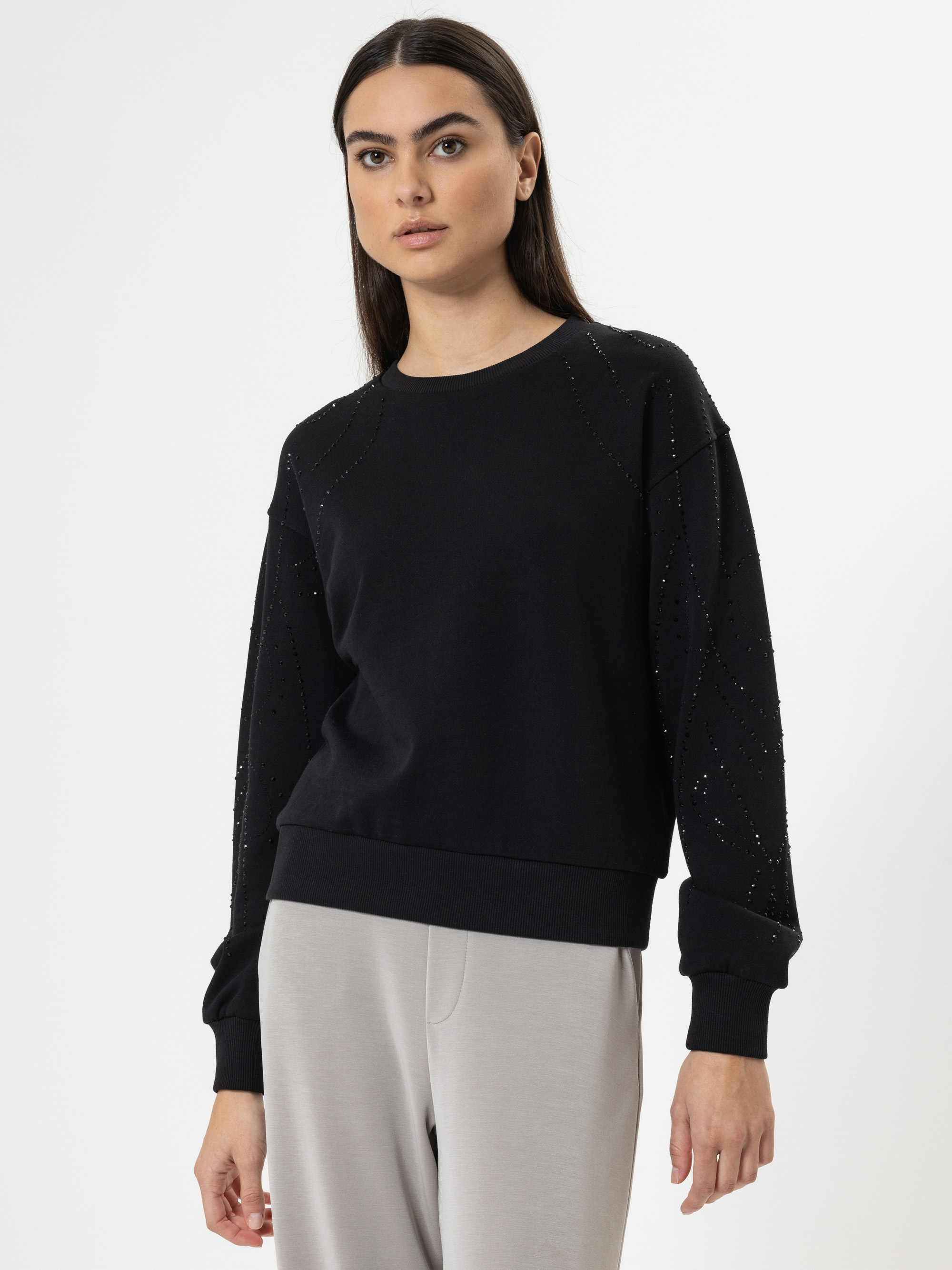 Eine Frau mit langen dunklen Haaren trägt ein schwarzes Sweatshirt mit dezenten Verzierungen und eine hellgraue Hose und steht vor einem schlichten weißen Hintergrund.