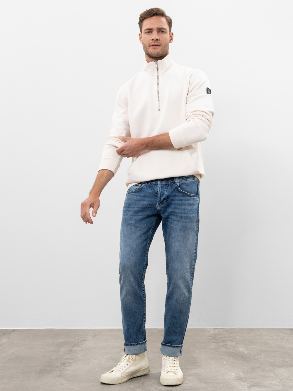 Ein Mann steht vor einem schlichten weißen Hintergrund und trägt ein helles Sweatshirt mit vierteljährlichem Reißverschluss, blaue Jeans und weiße Turnschuhe. Er hat einen Arm leicht erhoben und einen entspannten Gesichtsausdruck. Die Umgebung ist mi