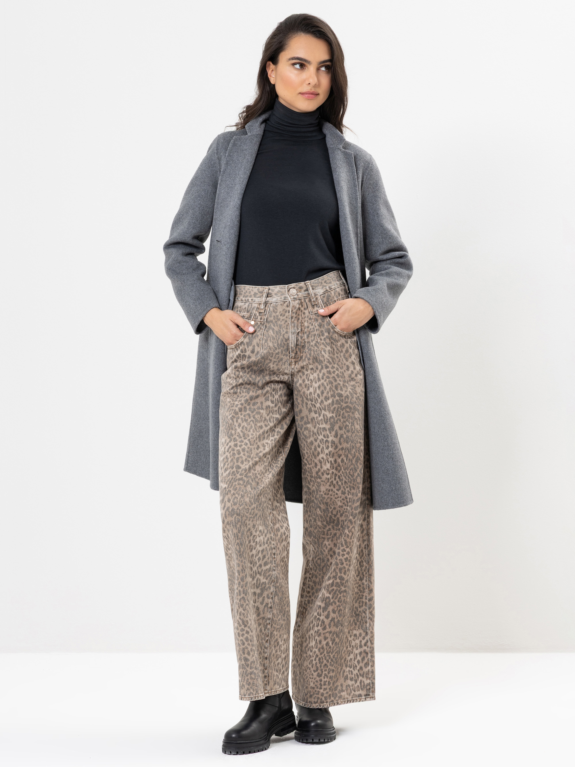 Eine Frau steht vor einem weißen Hintergrund, trägt einen grauen Mantel über einem schwarzen Rollkragenpullover, eine weit geschnittene Hose mit Leopardenmuster und schwarze Stiefel, hat die Hände in den Taschen und schaut leicht zur Seite.