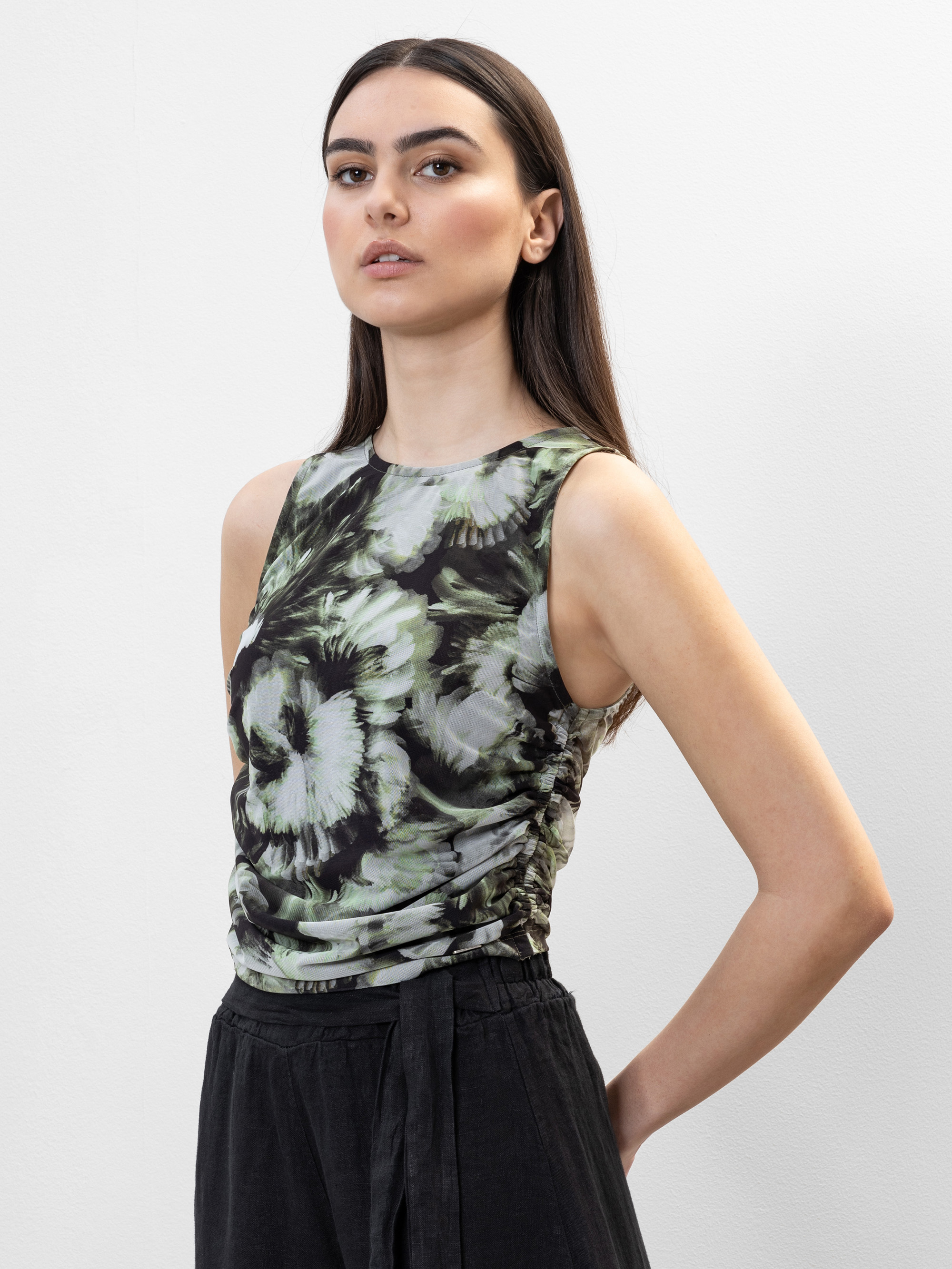 Eine Frau mit langen dunklen Haaren trägt ein ärmelloses, tailliertes Oberteil mit grünem und schwarzem Blumenmuster und eine schwarze Hose mit hoher Taille, die vor einem schlichten weißen Hintergrund steht.