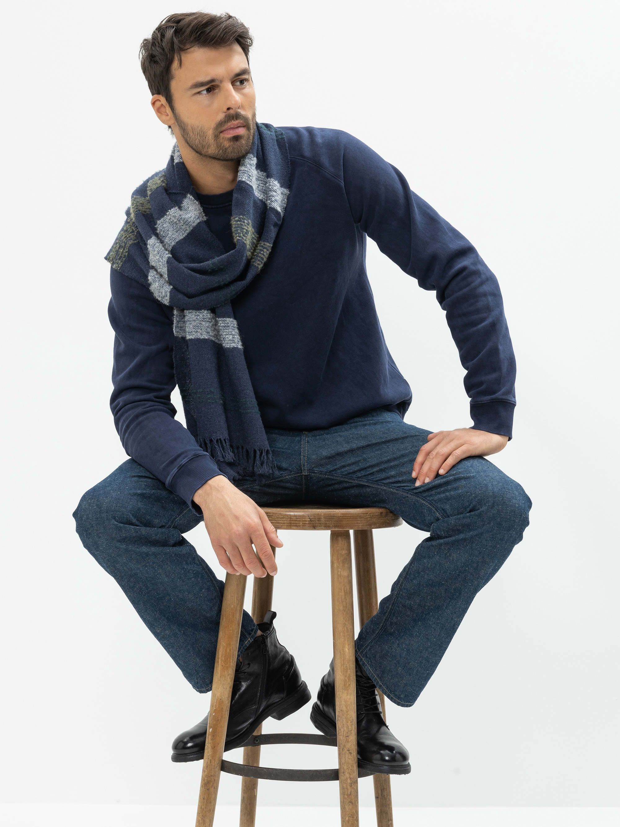 Ein Mann in einem marineblauen Pullover, blauen Jeans und schwarzen Stiefeln sitzt auf einem Holzschemel. Er hat sich einen blau-grau karierten Schal um den Hals gelegt und blickt zur Seite vor einem schlichten weißen Hintergrund.