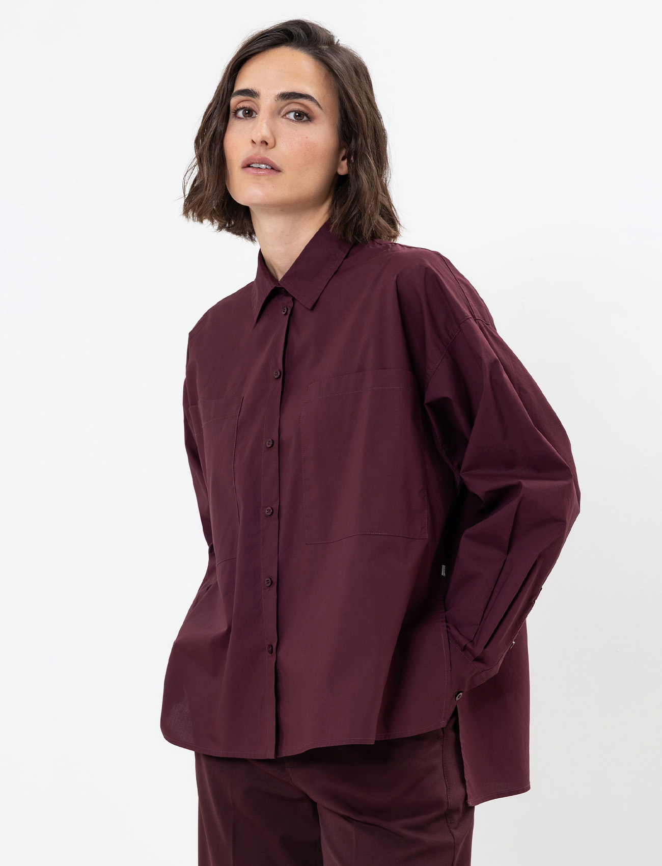 Eine Frau mit schulterlangen braunen Haaren trägt ein locker sitzendes, langärmeliges kastanienbraunes Button-up-Hemd und eine dazu passende Hose und steht vor einem schlichten weißen Hintergrund.