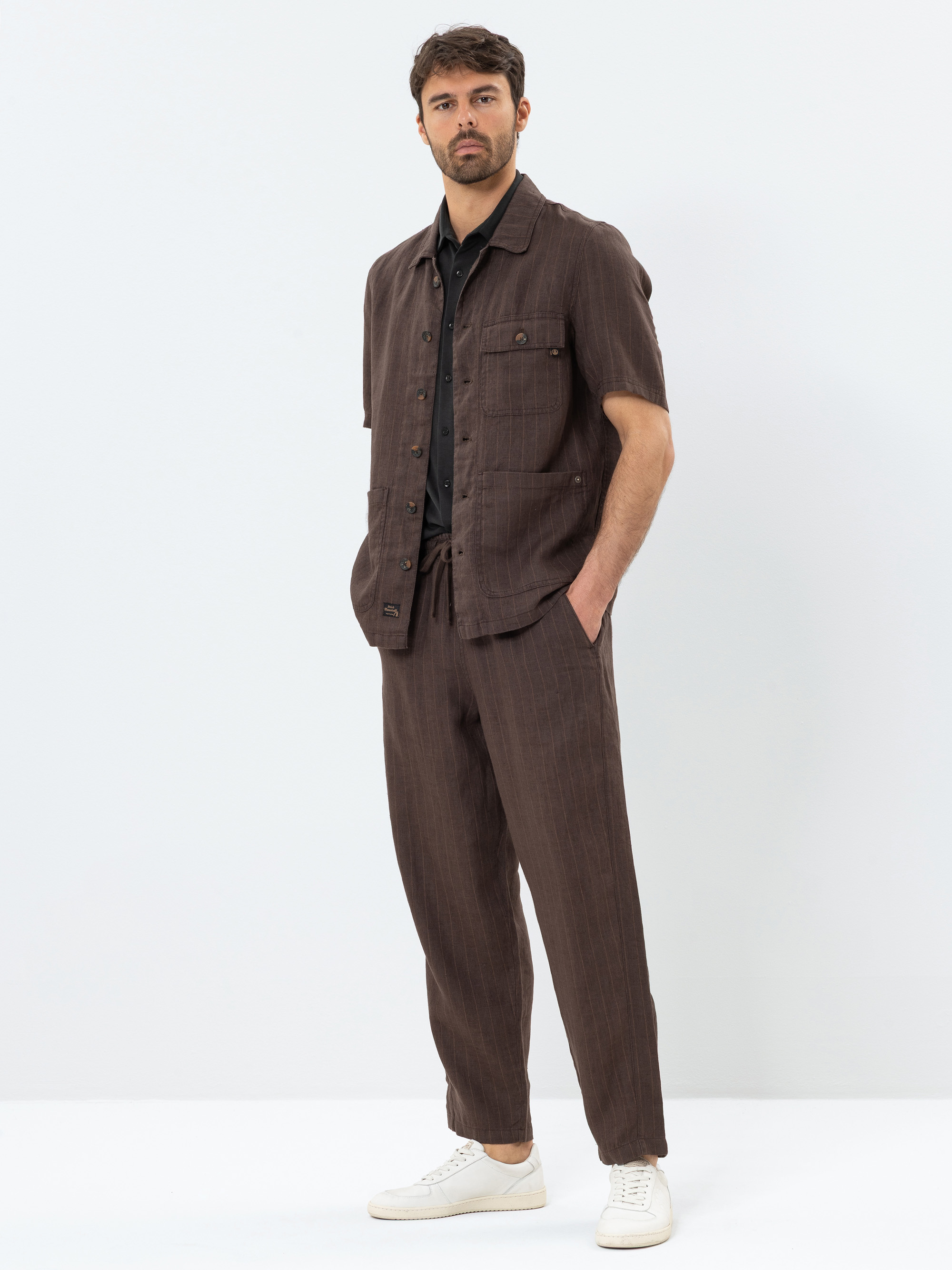 Ein Mann steht vor einem schlichten, hellen Hintergrund und trägt ein braunes, kurzärmeliges Button-up-Hemd mit passender Hose, darunter ein schwarzes Hemd und weiße Turnschuhe. Er hat eine Hand in der Tasche und einen neutralen Gesichtsausdruck.