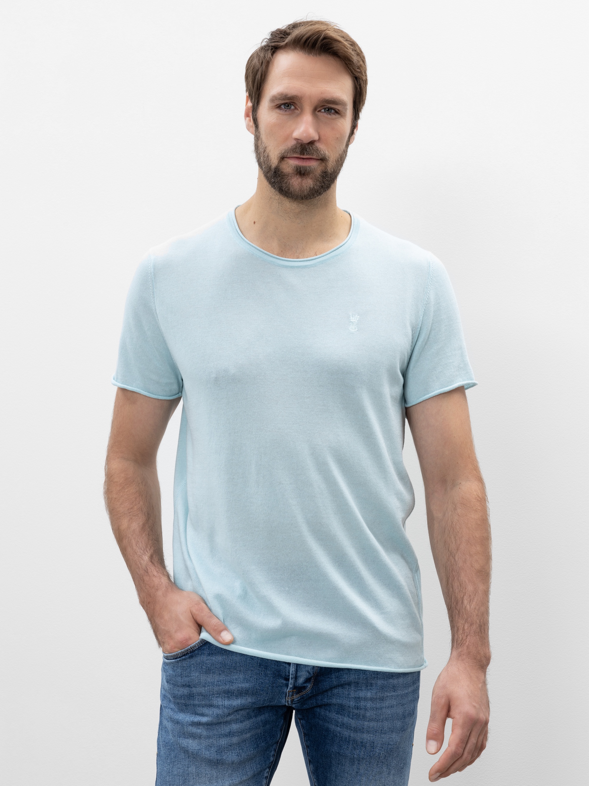 Ein Mann mit kurzen braunen Haaren und einem Bart, der ein hellblaues T-Shirt und eine blaue Jeans trägt, steht vor einem schlichten weißen Hintergrund, eine Hand in der Tasche und mit neutraler Miene.