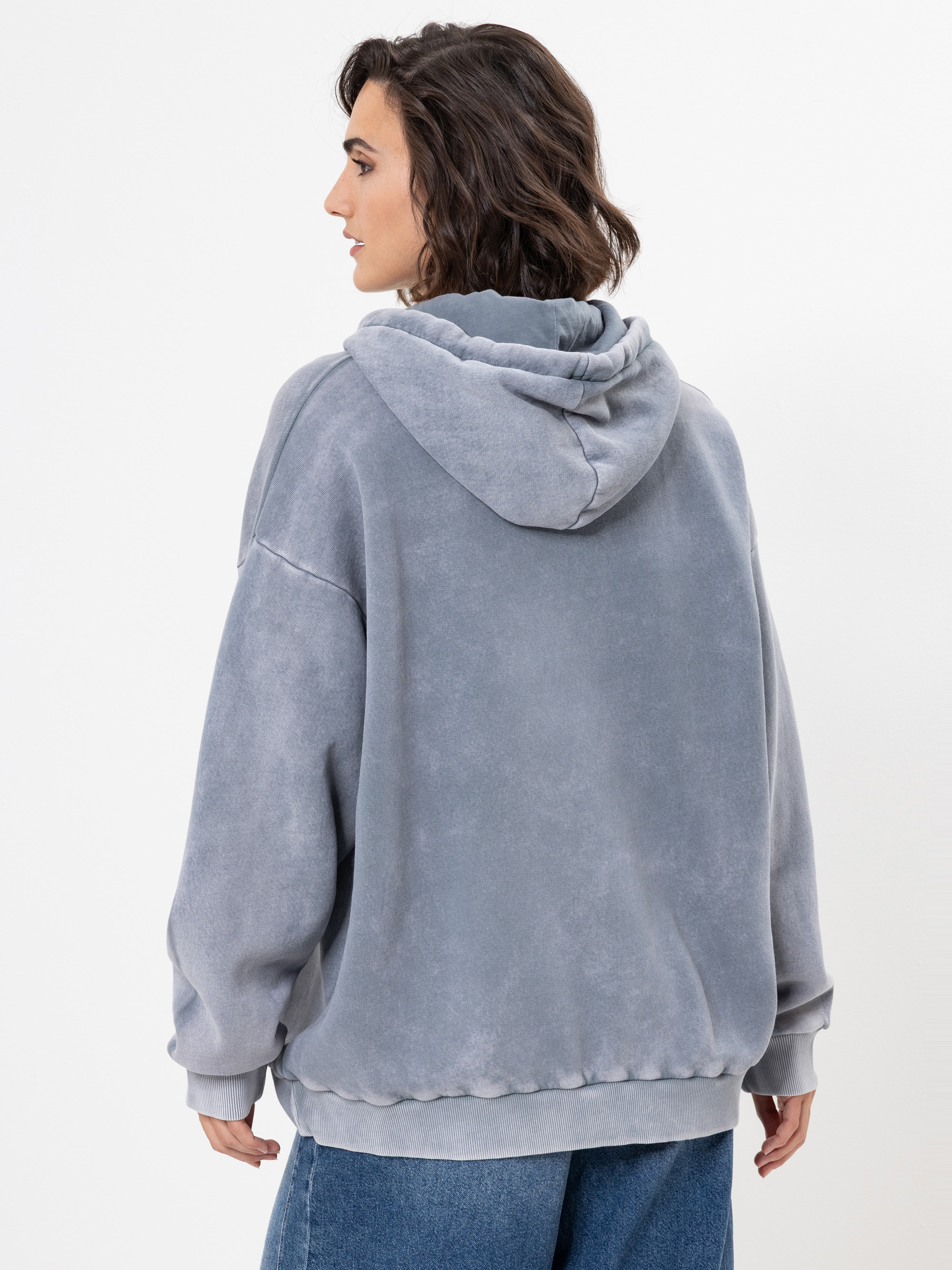 Eine Person mit gewelltem braunem Haar, bekleidet mit einem lockeren hellgrauen Kapuzenpulli und blauen Jeans, steht mit dem Gesicht zur Seite vor einem schlichten weißen Hintergrund.