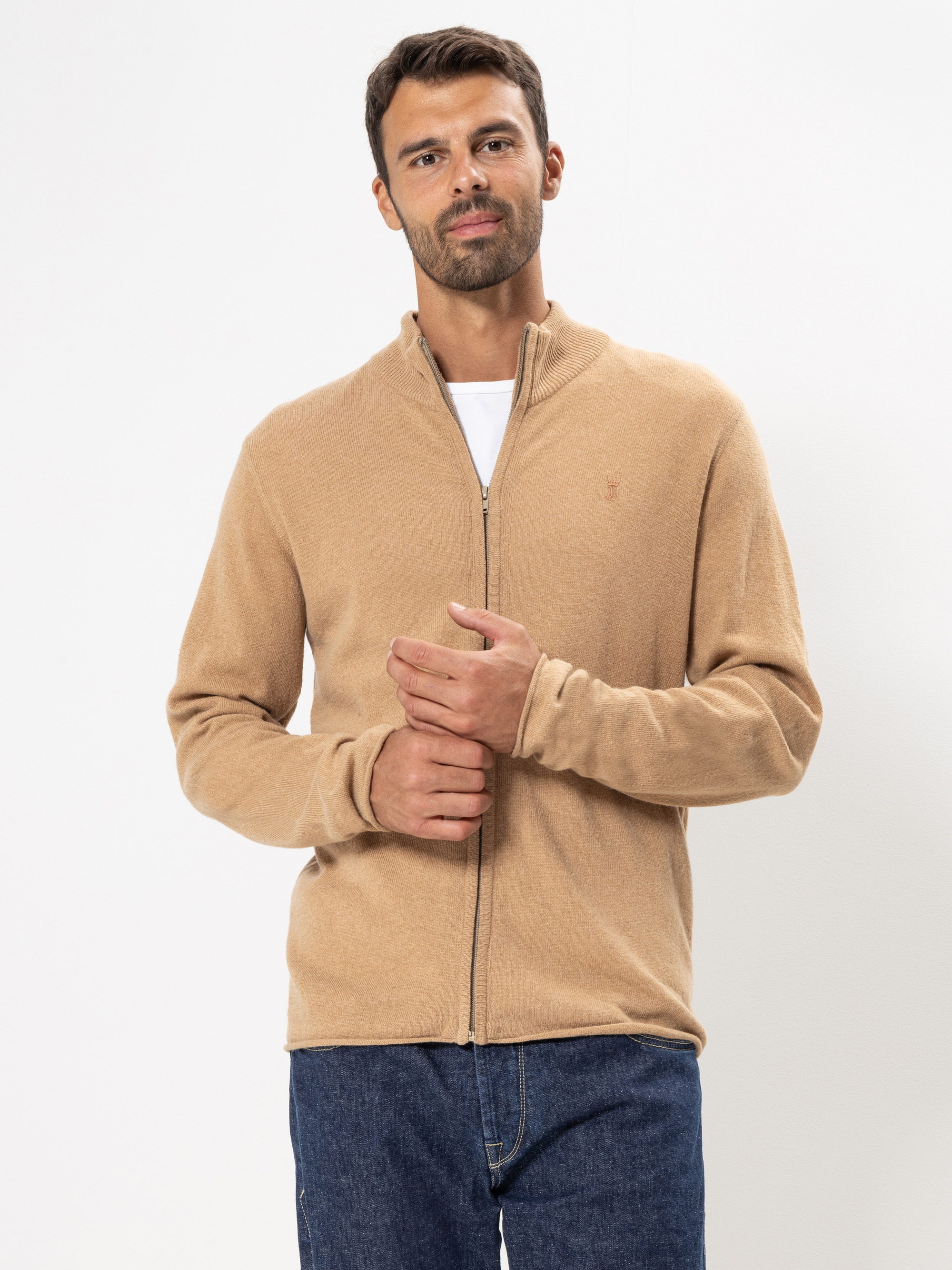 Ein Mann mit kurzen dunklen Haaren und einem Bart trägt einen hellbraunen Pullover mit Reißverschluss über einem weißen Hemd und blaue Jeans und steht vor einem schlichten weißen Hintergrund.