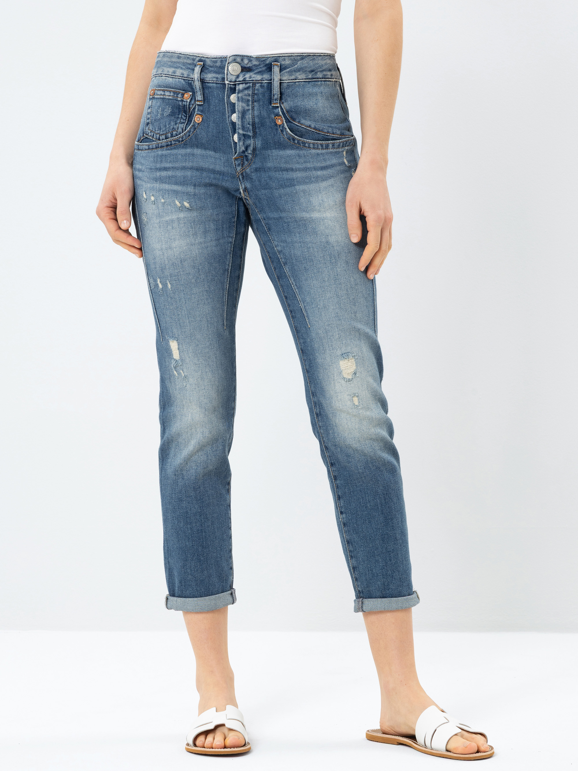 Eine Person, die eine blaue Jeans mit Manschetten, ein weißes ärmelloses Oberteil und weiße Sandalen trägt, steht vor einem schlichten hellen Hintergrund. Ihre Hände ruhen an ihren Seiten.