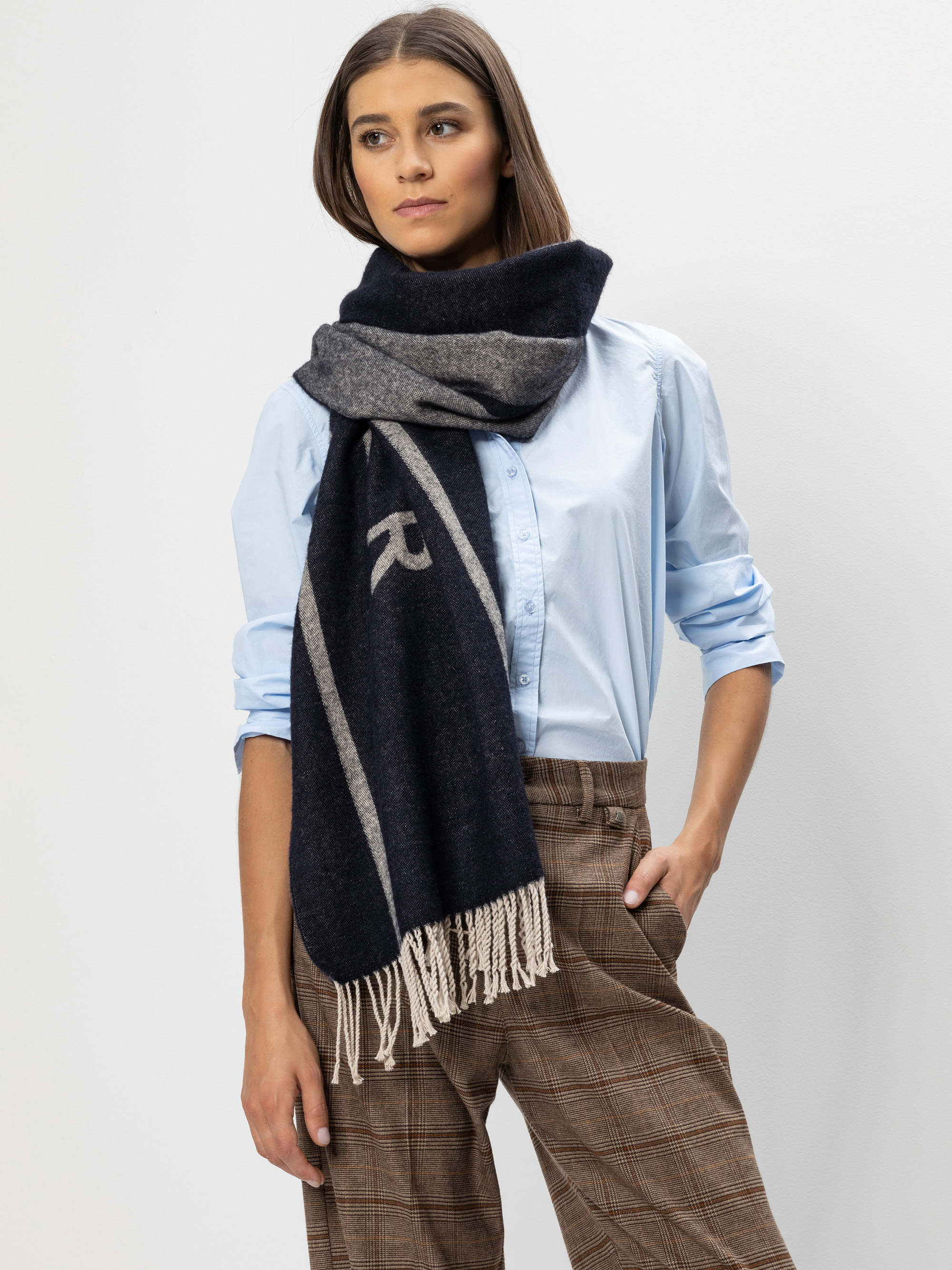 Eine Frau, die ein hellblaues Button-up-Hemd, eine karierte braune Hose und einen großen marineblauen und grauen Schal mit Fransen trägt, steht vor einem schlichten weißen Hintergrund und hat eine Hand in der Tasche.