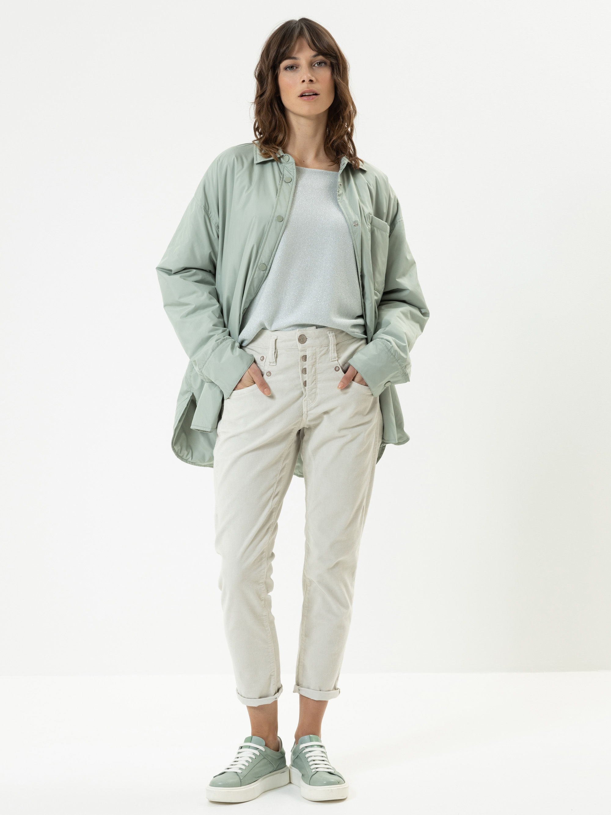 Eine Frau steht vor einem schlichten weißen Hintergrund und trägt ein hellgrünes, übergroßes Button-up-Hemd über einem hellblauen Oberteil, eine weiße Hose mit hoher Taille und passende grüne Turnschuhe. Sie hat die Hände in den Taschen und blick