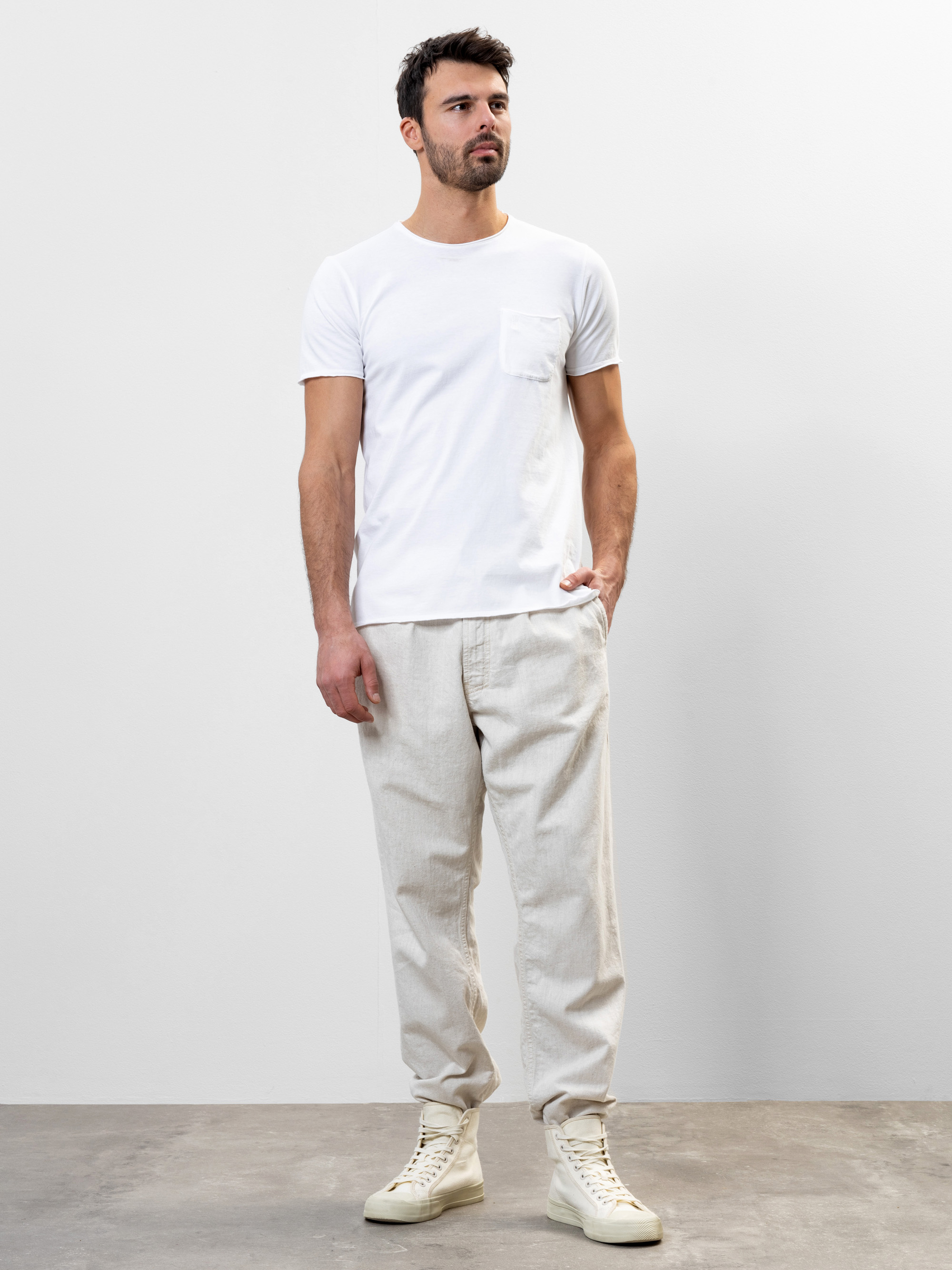 Ein Mann steht vor einer schlichten weißen Wand. Er trägt ein weißes, kurzärmeliges T-Shirt mit einer Brusttasche, eine hellbeige Hose und cremefarbene, hohe Turnschuhe. Er hat kurzes dunkles Haar und einen Bart und blickt leicht zur Seite.