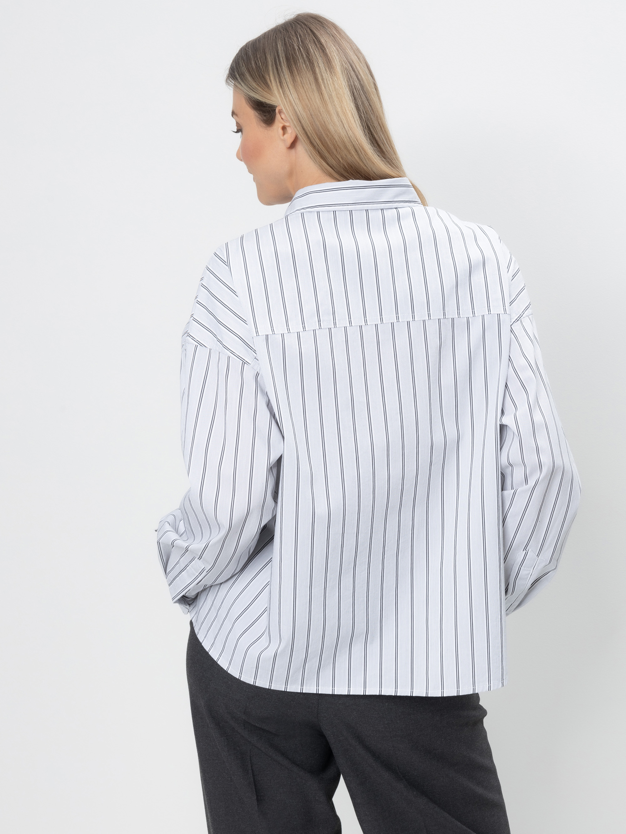 Eine Frau mit glattem hellbraunem Haar steht mit dem Rücken zur Kamera. Sie trägt ein weißes Button-up-Hemd mit vertikalen schwarzen Streifen und eine dunkle Hose vor einem schlichten weißen Hintergrund.