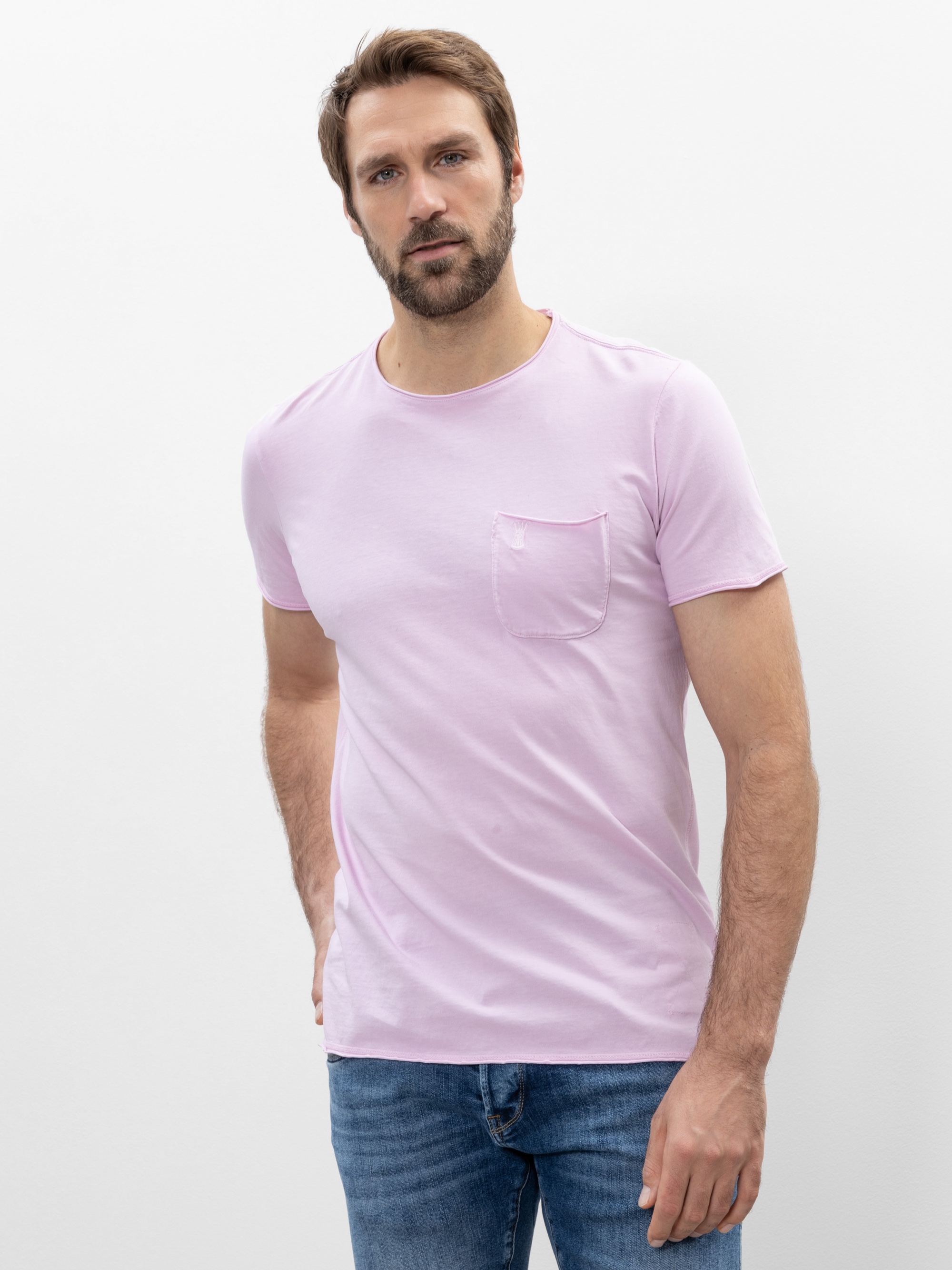 Ein Mann mit hellbraunem Haar und Bart trägt ein hellrosa T-Shirt mit Brusttasche und eine blaue Jeans und steht vor einem schlichten weißen Hintergrund.