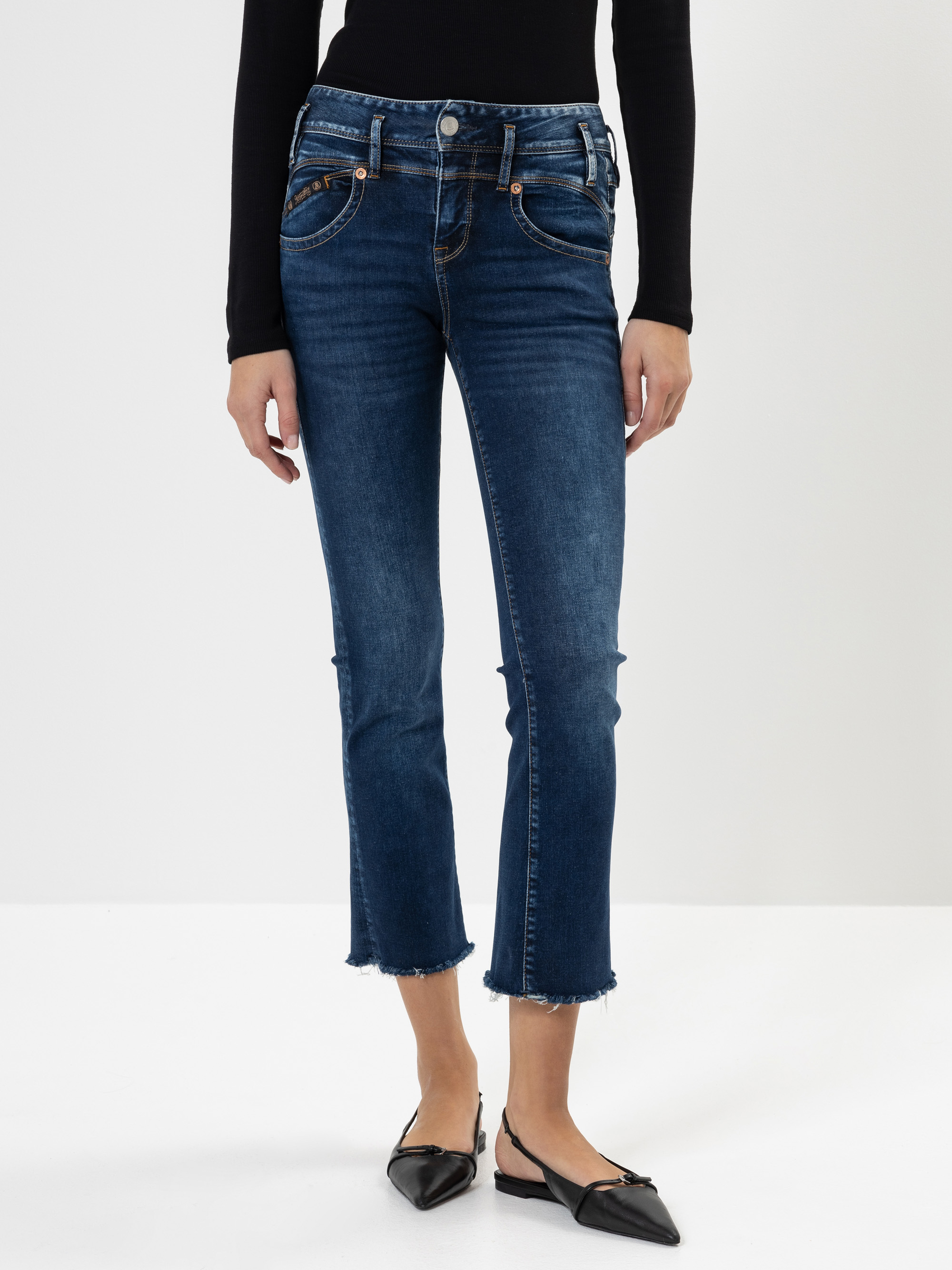Eine Person, die eine dunkelblaue, knöchellange Jeans mit ausgefranstem Saum, ein schwarzes, langärmeliges Oberteil und schwarze, spitz zulaufende Slingback-Flats trägt, steht vor einem schlichten weißen Hintergrund.