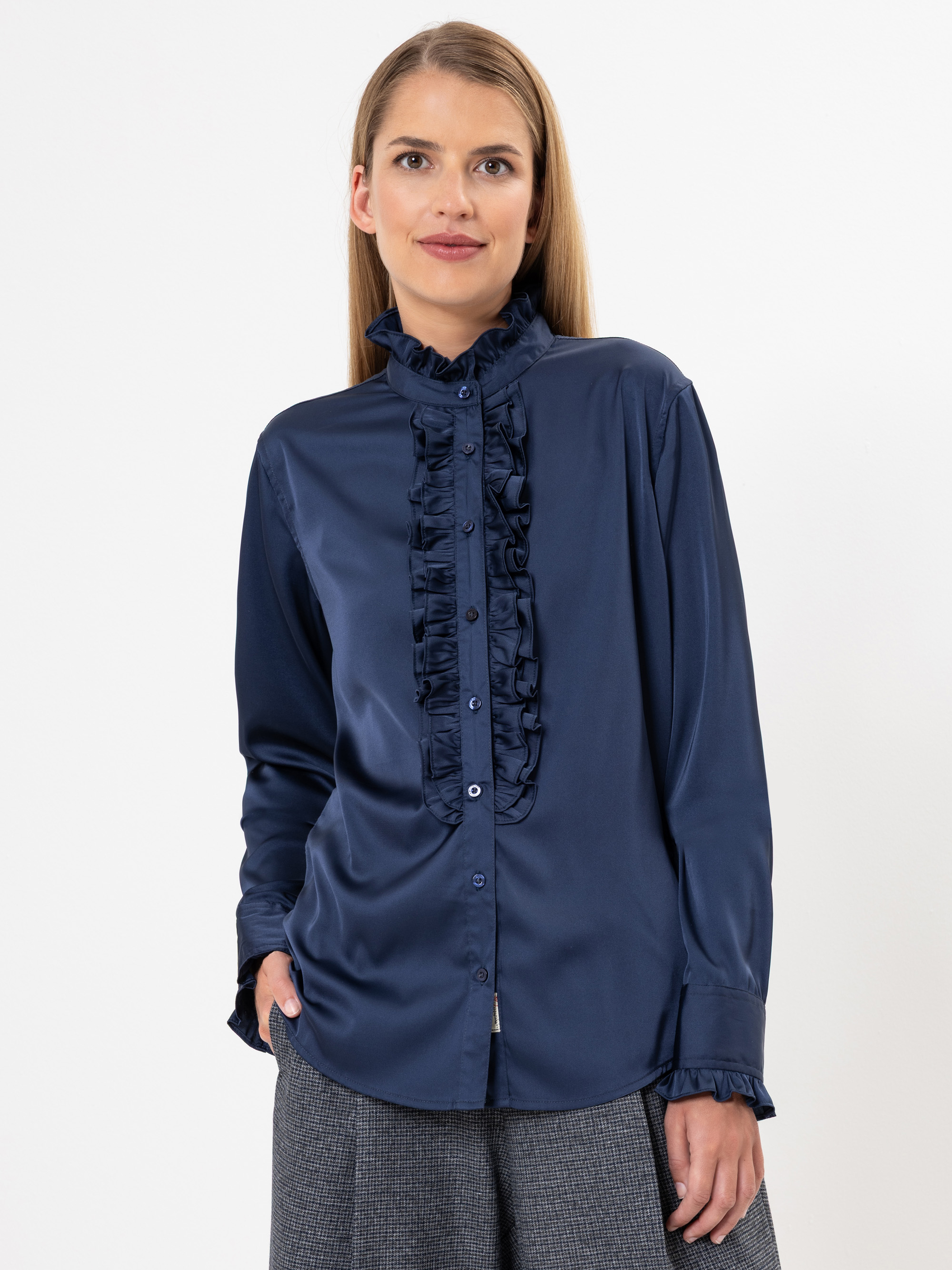 Eine Frau mit langem, glattem blondem Haar trägt eine marineblaue, langärmelige Bluse mit Rüschenbesatz auf der Vorderseite und an den Manschetten. Sie steht vor einem schlichten weißen Hintergrund, blickt leicht nach links und lächelt sanft.