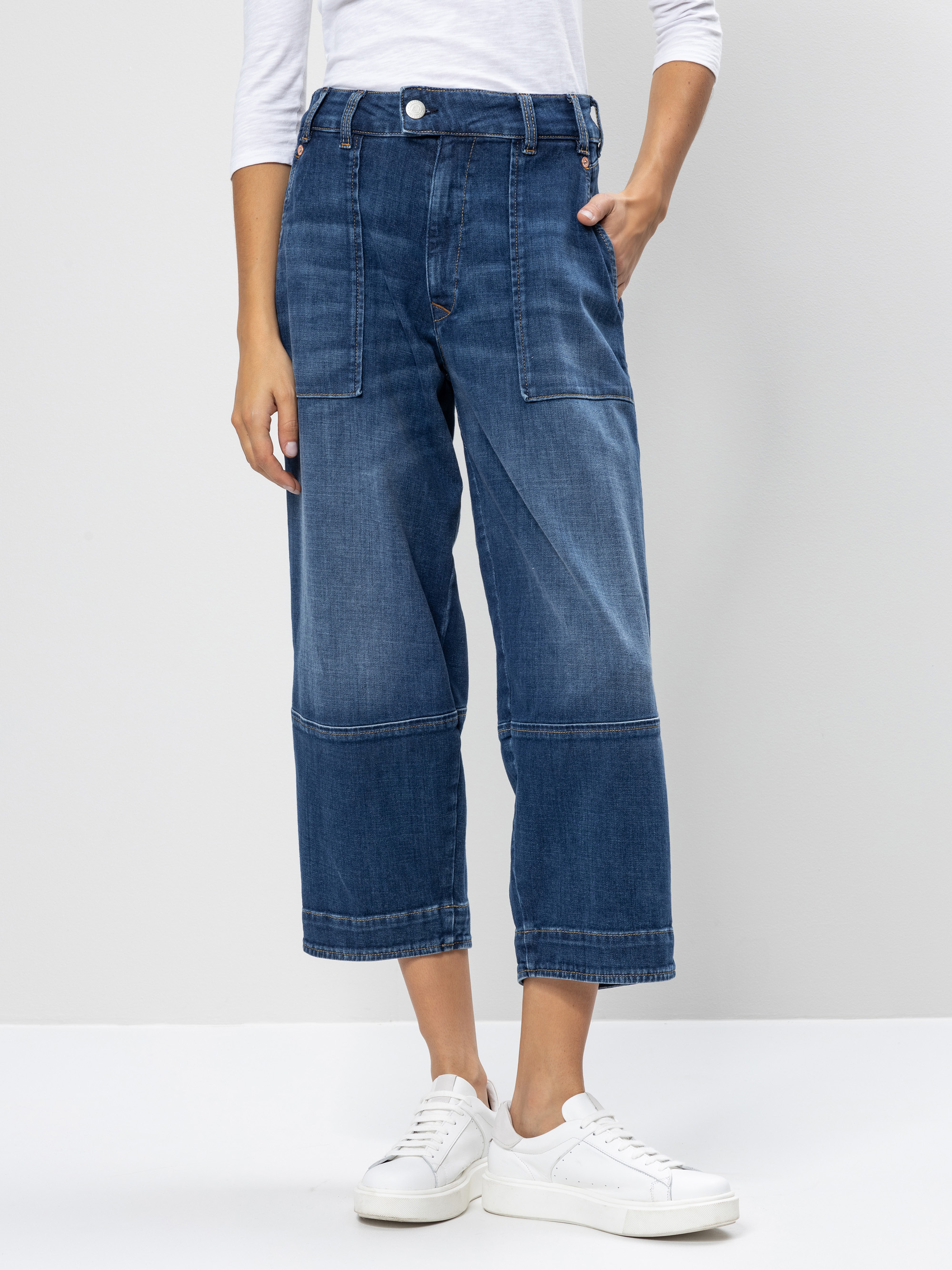 Eine Person, die ein weißes, langärmeliges Oberteil, eine blaue, weit geschnittene Jeans mit aufgesetzten Taschen und weiße Turnschuhe trägt, steht vor einem schlichten, hellen Hintergrund.