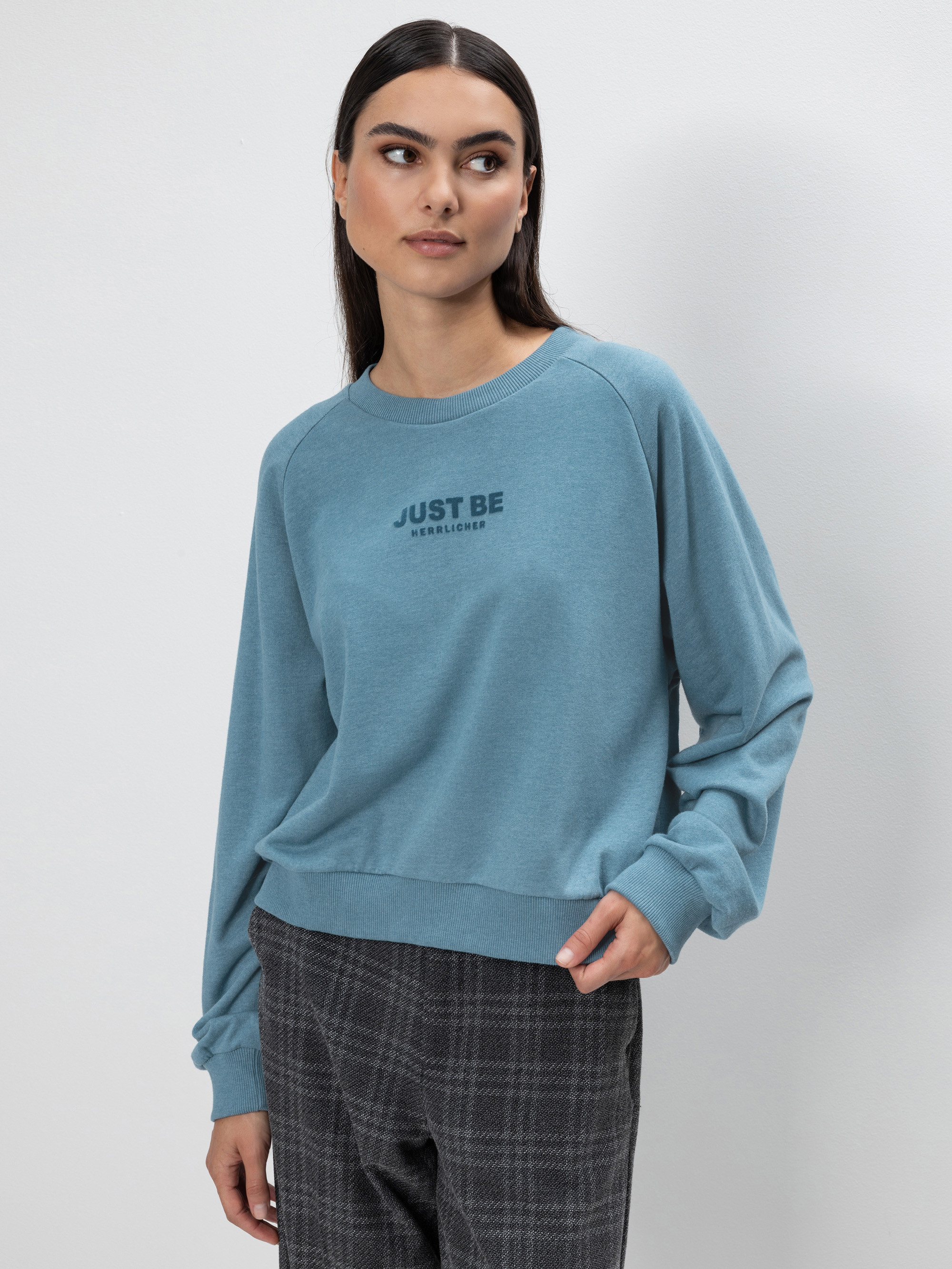 Eine Frau mit langen dunklen Haaren trägt ein hellblaues Sweatshirt mit der Aufschrift JUST BE und eine grau karierte Hose. Sie steht vor einem schlichten weißen Hintergrund und blickt mit einem neutralen Ausdruck leicht zur Seite.
