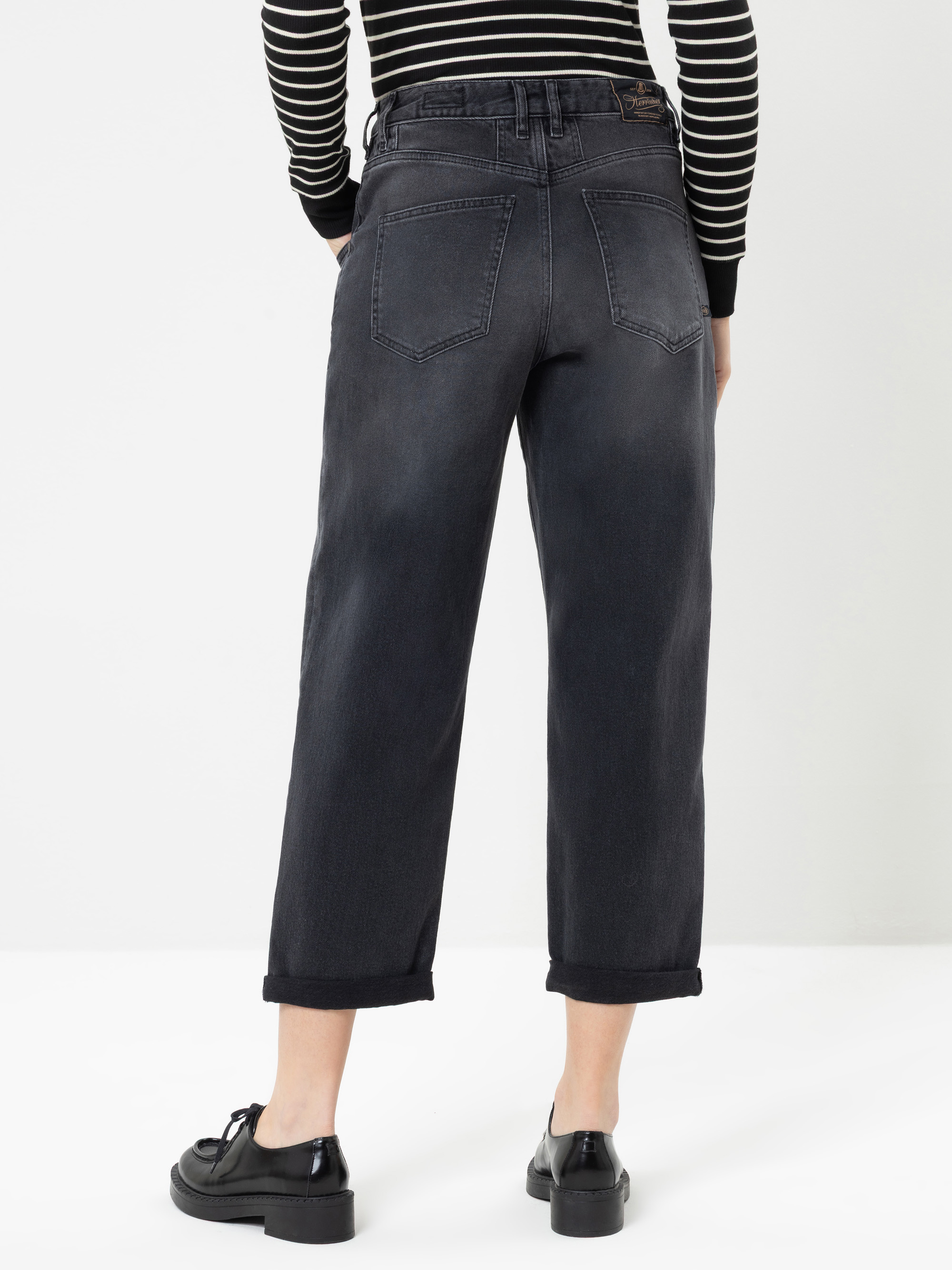 Eine Person, die eine schwarze, hoch taillierte Jeans mit Umschlag und ein schwarz-weiß gestreiftes Langarm-Oberteil trägt, steht mit dem Rücken zur Kamera vor einem weißen Hintergrund. Schwarze Schuhe runden das Outfit ab.