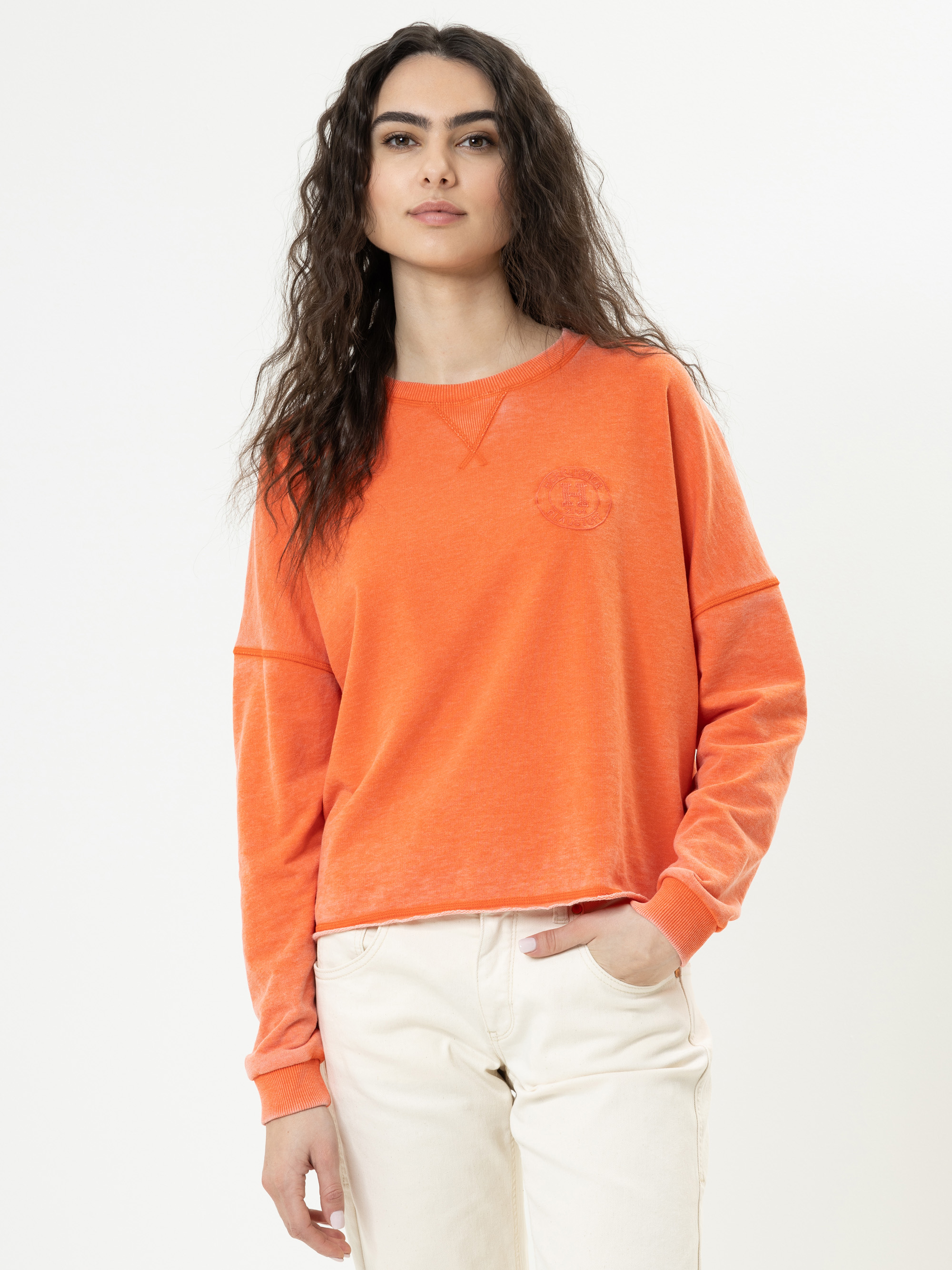 Eine Frau mit langen, gewellten dunklen Haaren steht vor einem einfarbig weißen Hintergrund und trägt ein orangefarbenes, langärmeliges Sweatshirt und eine cremefarbene Hose. Sie hat eine Hand in der Tasche und einen neutralen Gesichtsausdruck.