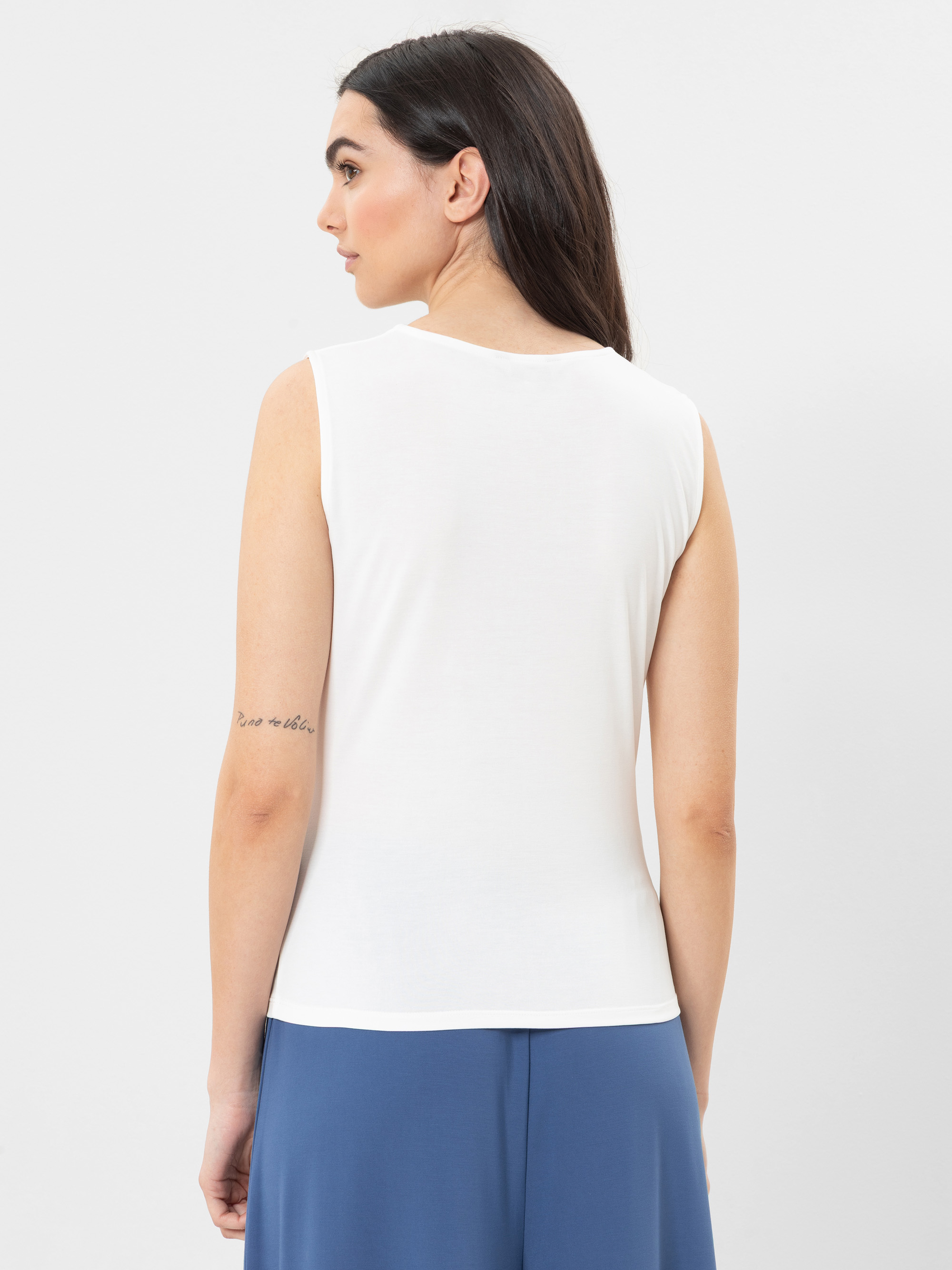 Eine Frau mit langen dunklen Haaren, die ein ärmelloses weißes Oberteil und eine blaue Hose trägt, steht von der Kamera abgewandt. Sie hat eine kleine Tätowierung auf ihrem linken Oberarm und posiert vor einem schlichten weißen Hintergrund.