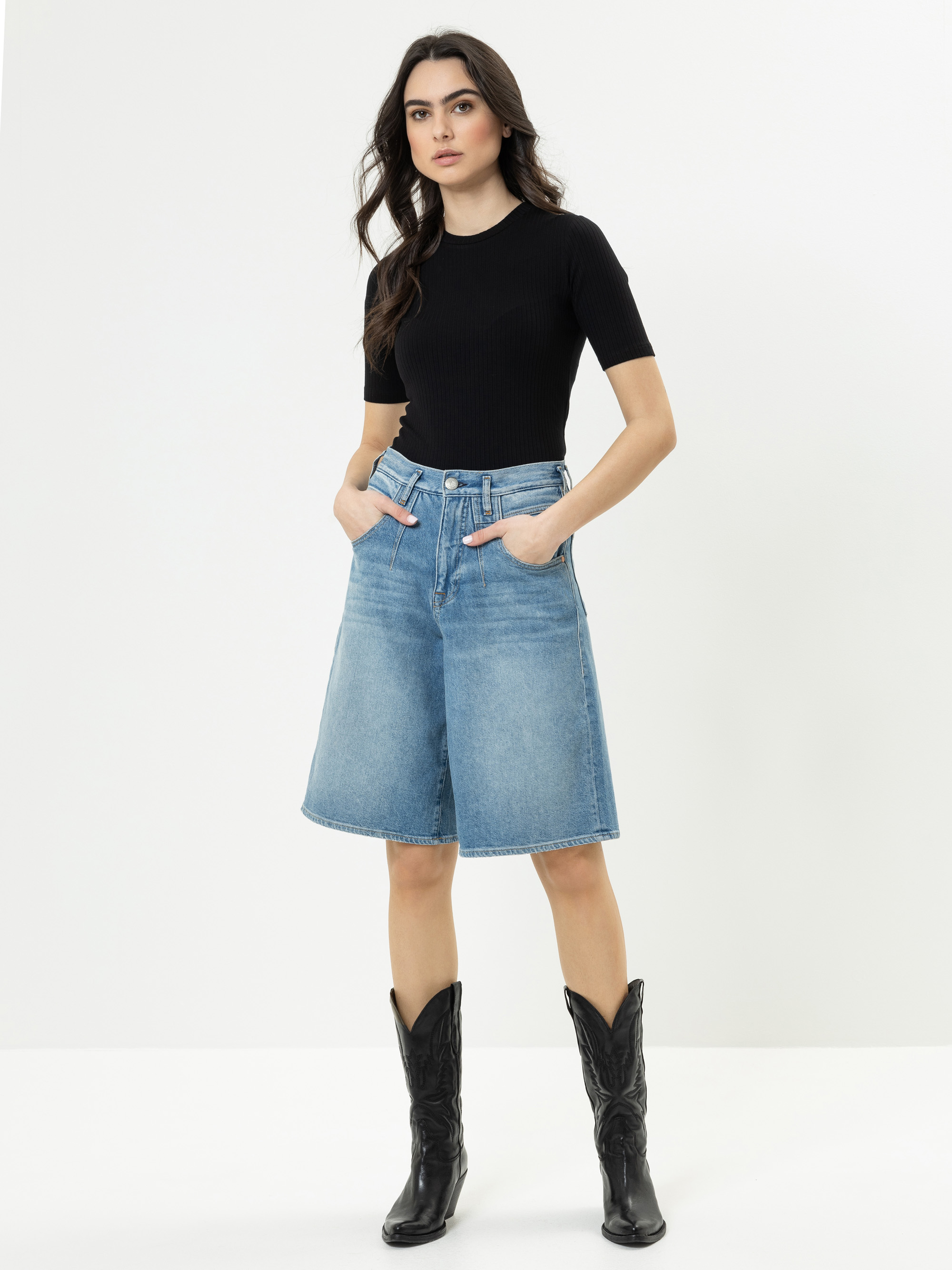 Eine Frau steht vor einem schlichten weißen Hintergrund, trägt ein schwarzes, kurzärmeliges Oberteil, einen knielangen Jeansrock und schwarze Cowboystiefel, die Hände in den Rocktaschen.