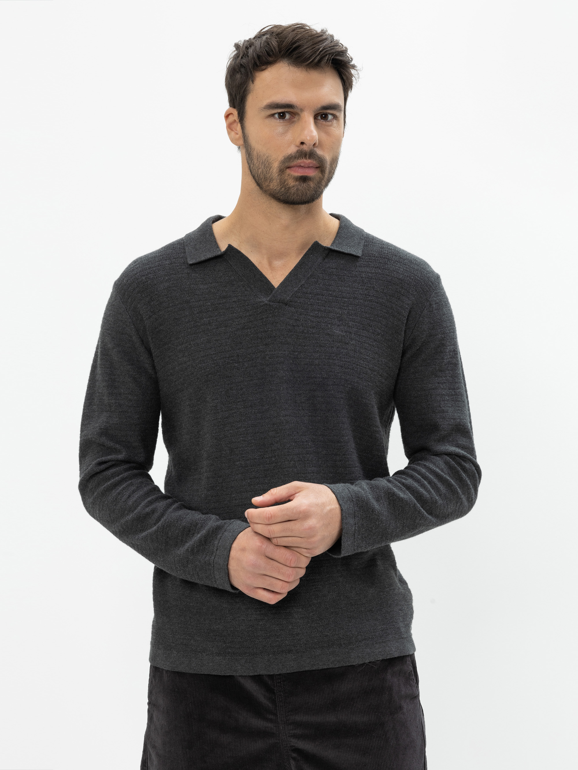 Ein Mann mit kurzen dunklen Haaren und einem Bart trägt einen dunklen langärmeligen Pullover mit einem V-förmigen Kragen. Er steht vor einem schlichten weißen Hintergrund und blickt mit locker gefalteten Händen leicht zur Seite.