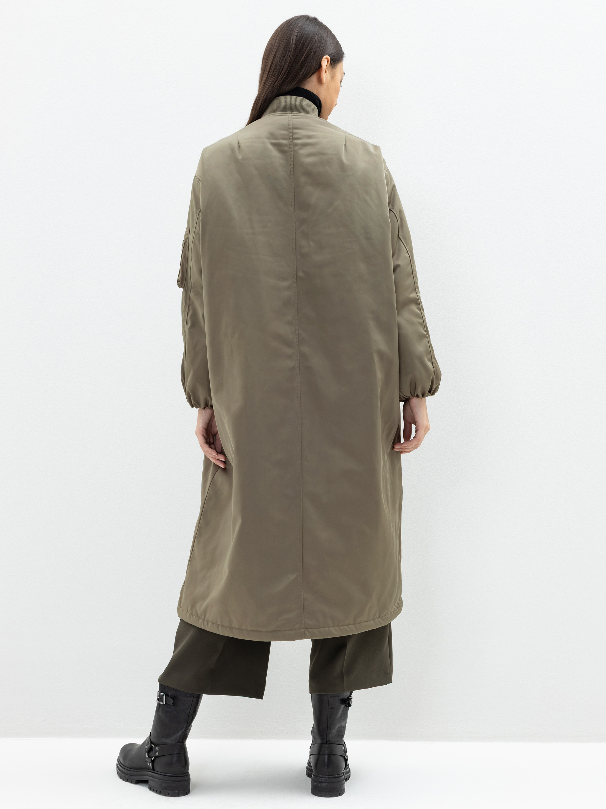 Eine Person mit langen dunklen Haaren, die einen olivgrünen langen Mantel, weite Hosen und schwarze Stiefel trägt, steht mit dem Gesicht von der Kamera weg vor einem schlichten weißen Hintergrund.
