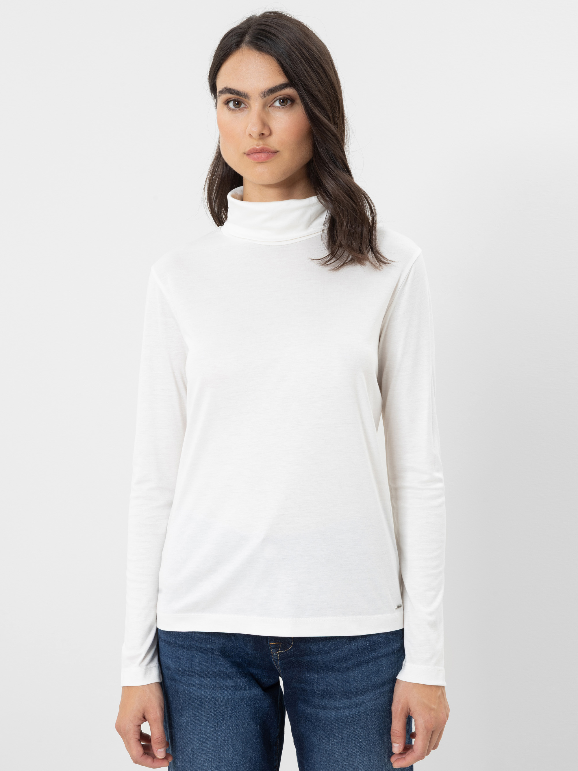 Eine Frau mit langen, dunklen Haaren trägt ein weißes Langarm-Rollkragen-Shirt und eine blaue Jeans und steht vor einem schlichten weißen Hintergrund.