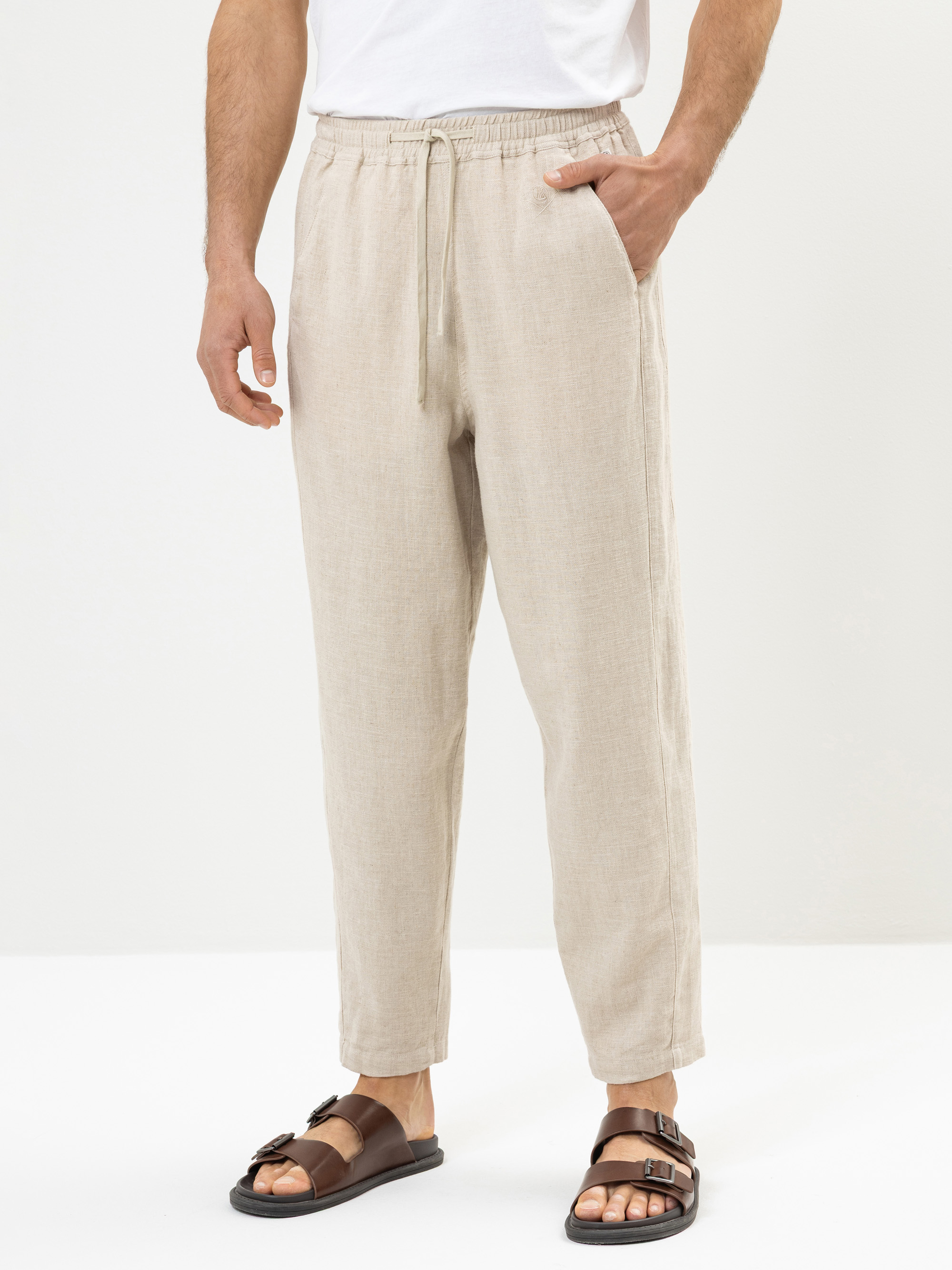Eine Person, die eine hellbeige Leinenhose, ein weißes T-Shirt und braune Sandalen trägt, steht vor einem schlichten weißen Hintergrund und hat eine Hand in der Tasche.
