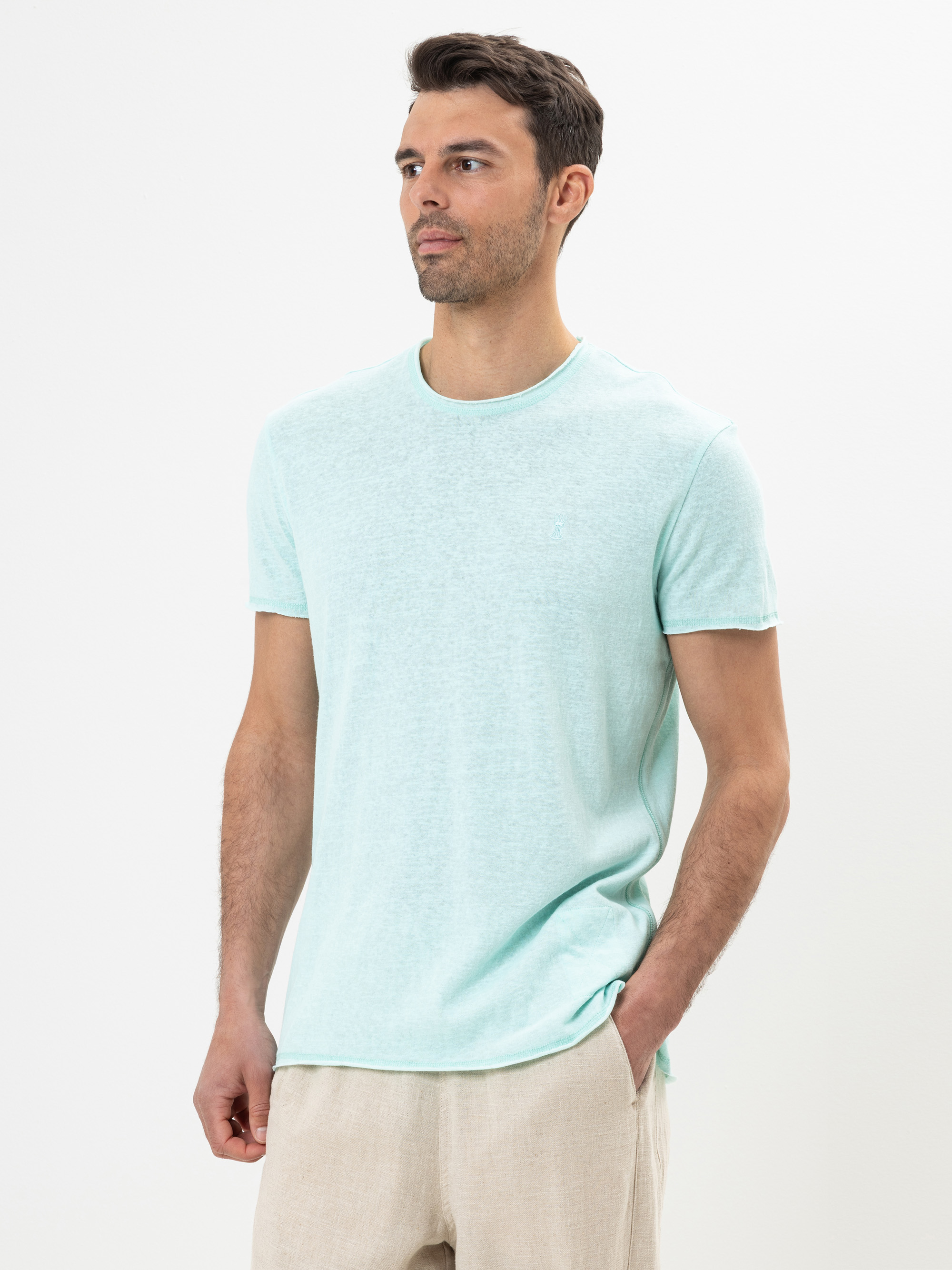 Ein Mann in einem hellen, mintgrünen, kurzärmeligen T-Shirt und einer beigen Hose steht vor einem schlichten weißen Hintergrund und schaut leicht zur Seite, eine Hand in der Tasche.