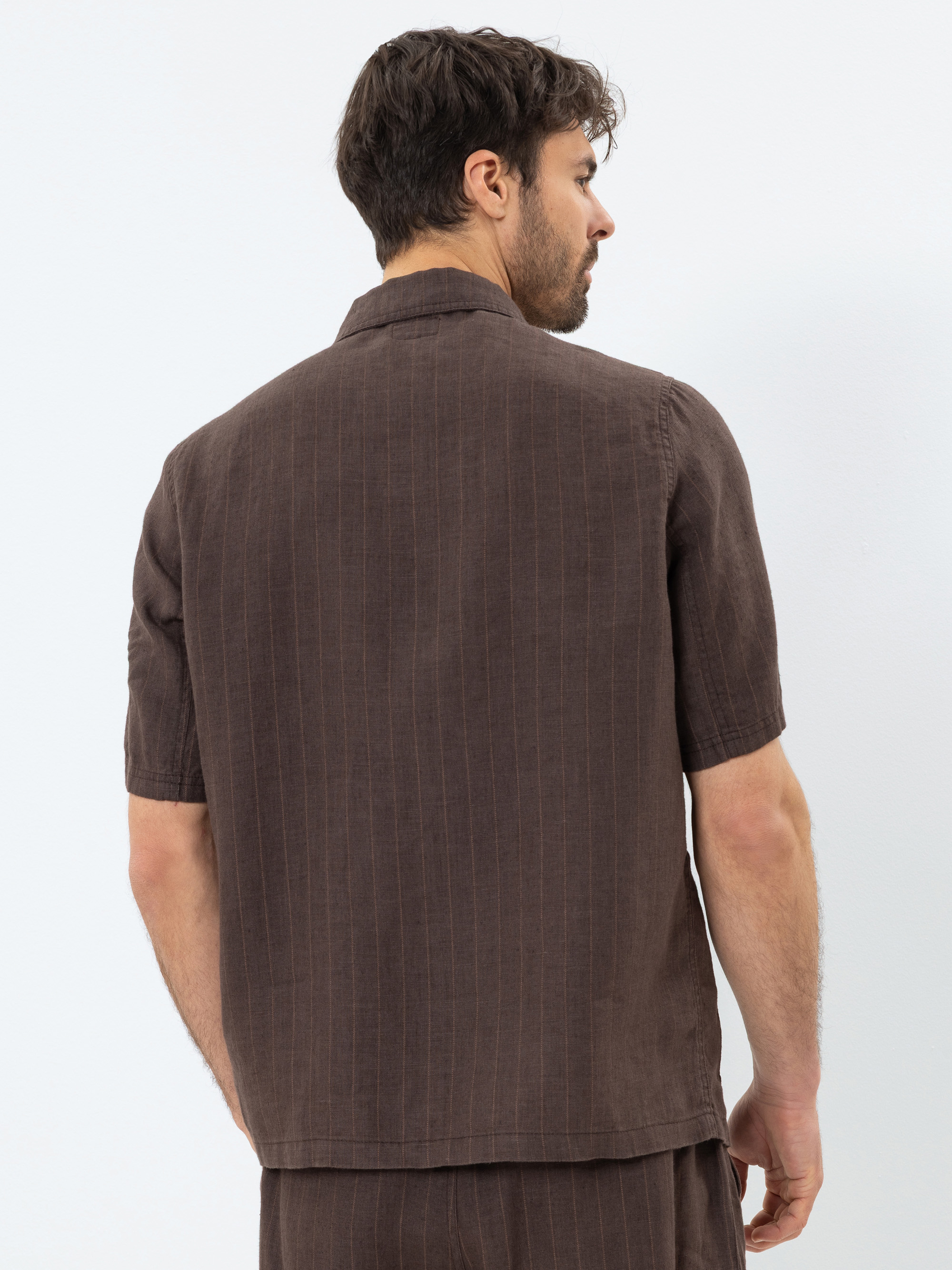 Ein Mann mit kurzen braunen Haaren, von hinten gesehen, trägt ein braunes, kurzärmeliges Hemd mit Kragen und dezenten vertikalen Nadelstreifen sowie eine dazu passende braune Hose und steht vor einem schlichten weißen Hintergrund.