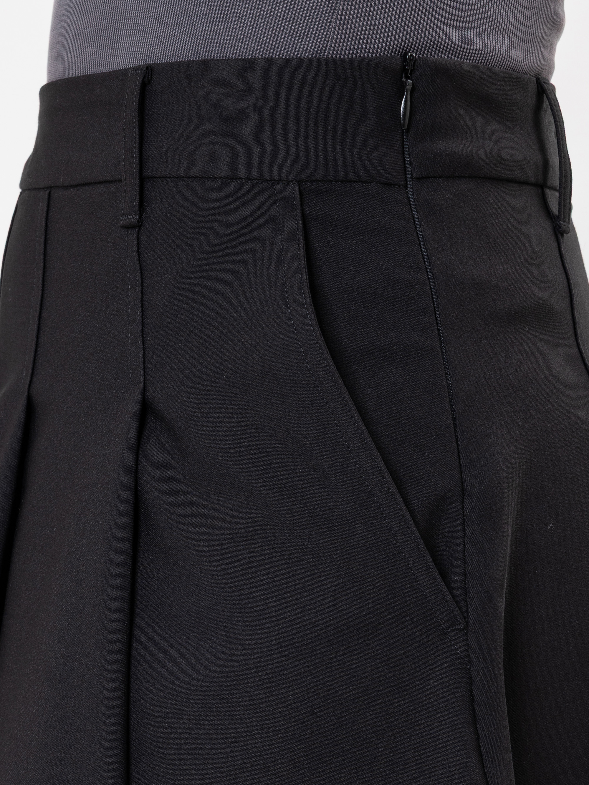 Nahaufnahme einer Person, die eine schwarze Bundfaltenhose mit einem seitlichen Reißverschluss und einer Tasche trägt, kombiniert mit einem grauen gerippten Oberteil.