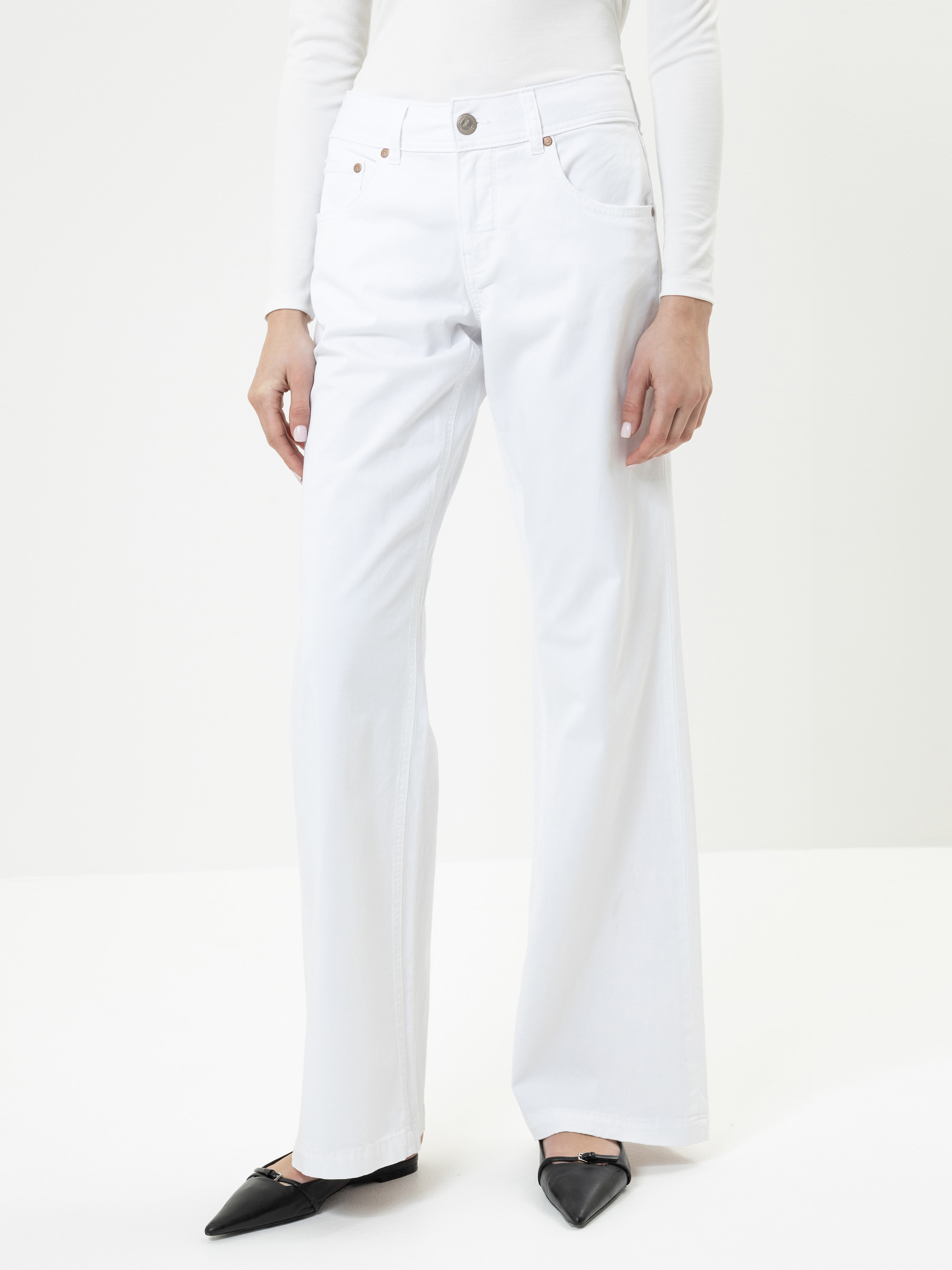 Eine Person, die ein weißes, langärmeliges Oberteil und eine weiße Jeans mit hoher Taille und weitem Bein trägt, dazu schwarze Schuhe mit spitzen Zehen, steht vor einem einfarbig weißen Hintergrund.