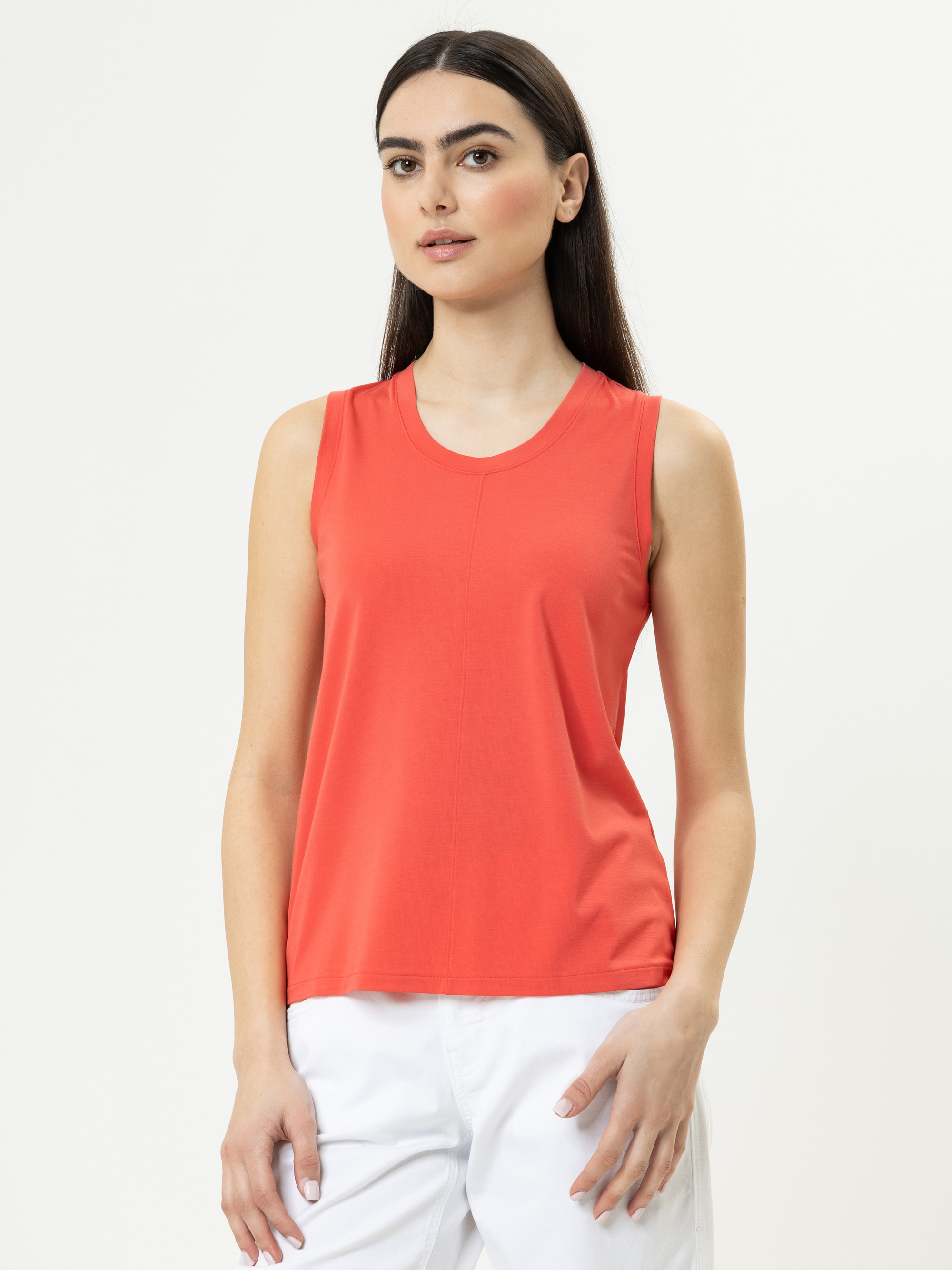 Eine Frau mit langen dunklen Haaren trägt ein ärmelloses, korallenrotes Oberteil und eine weiße Hose. Sie steht vor einem schlichten, hellen Hintergrund und blickt mit einem neutralen Ausdruck leicht zur Seite.