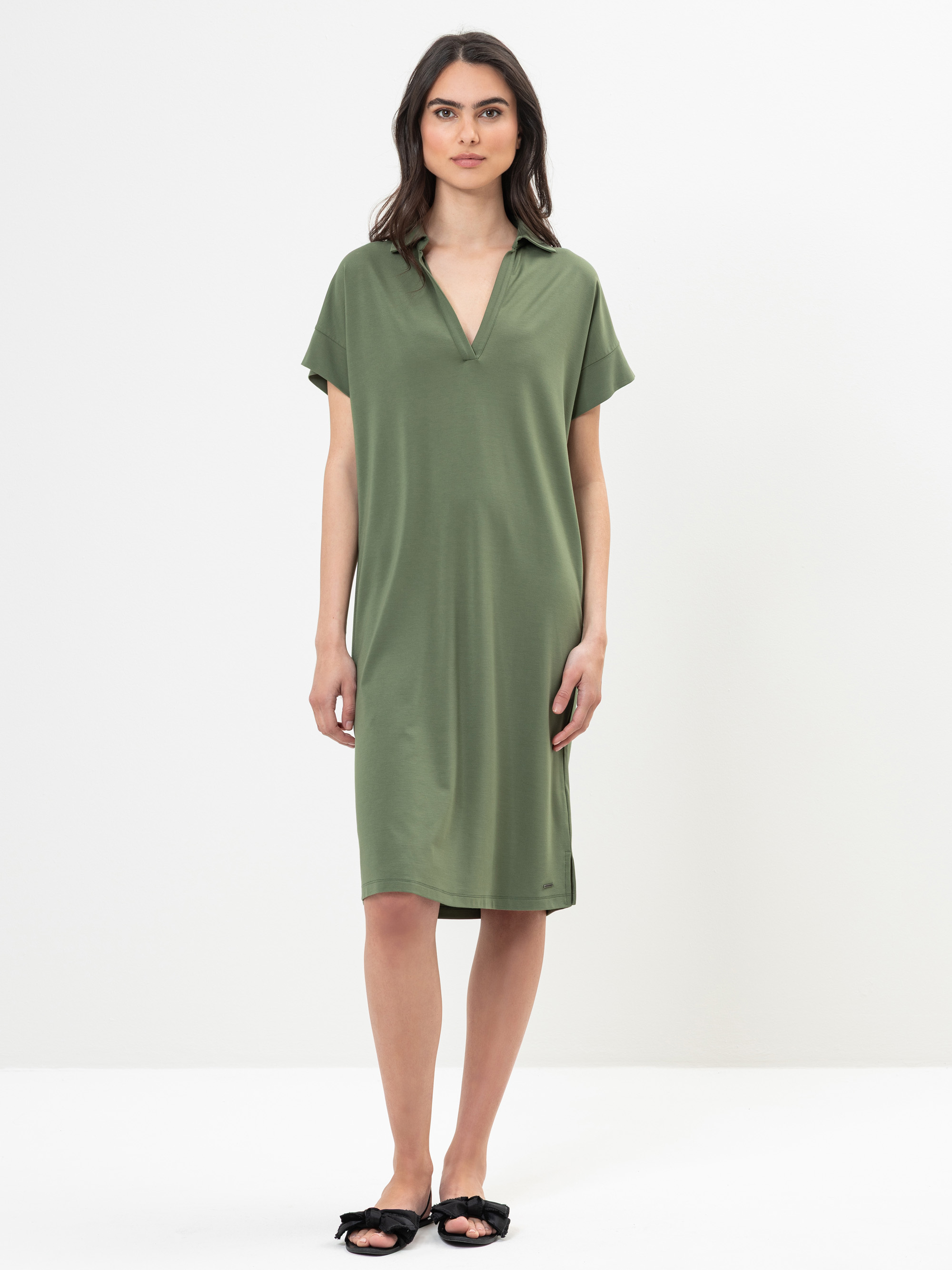 Eine Frau steht vor einem schlichten weißen Hintergrund und trägt ein locker sitzendes, knielanges olivgrünes Kleid mit kurzen Ärmeln und V-Ausschnitt. Sie trägt außerdem schwarze flache Slipper mit Schleifen.